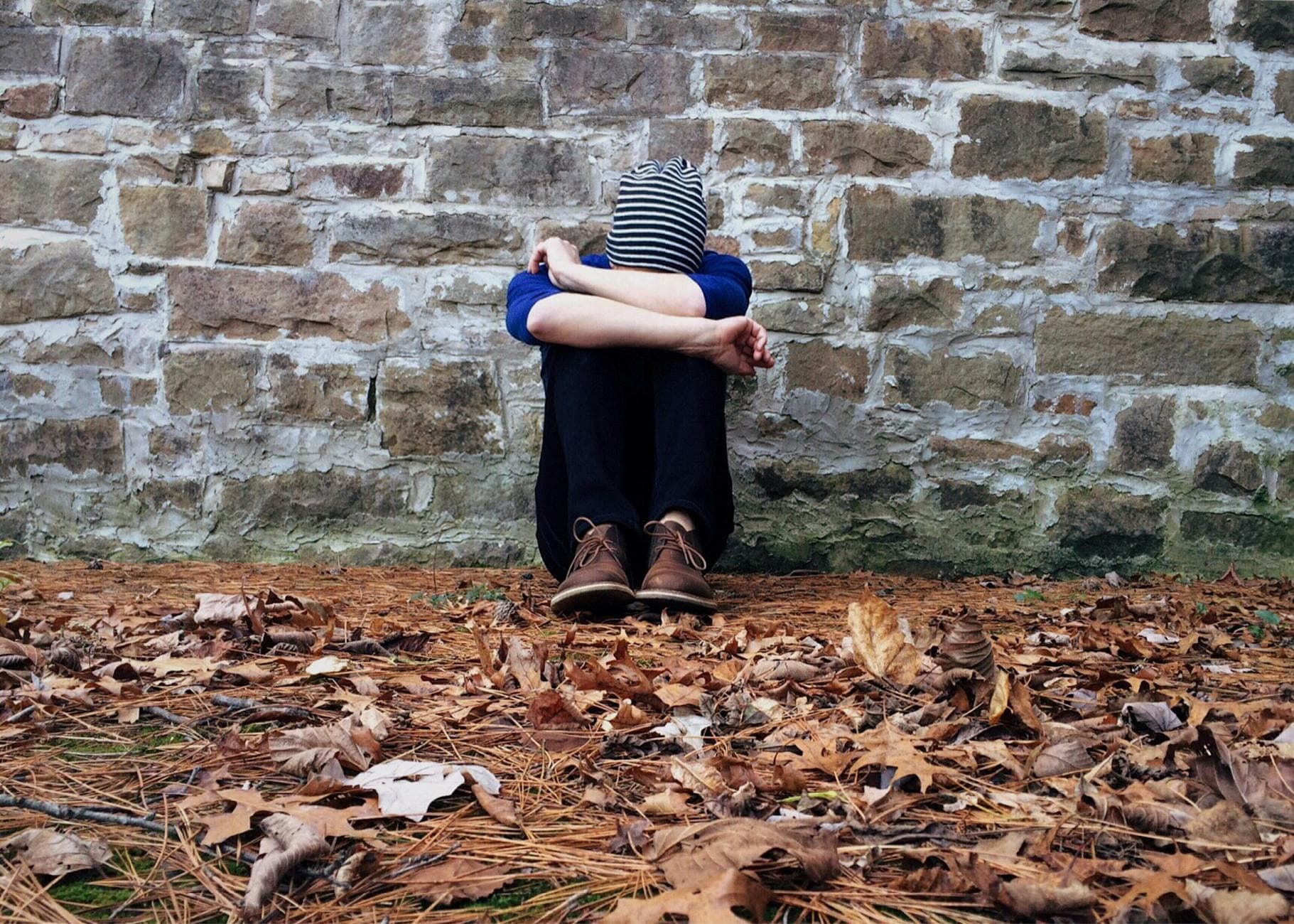 A solitary figure sits against a stone wall surrounded by fallen leaves, expressing isolation. - seasonal depression teens