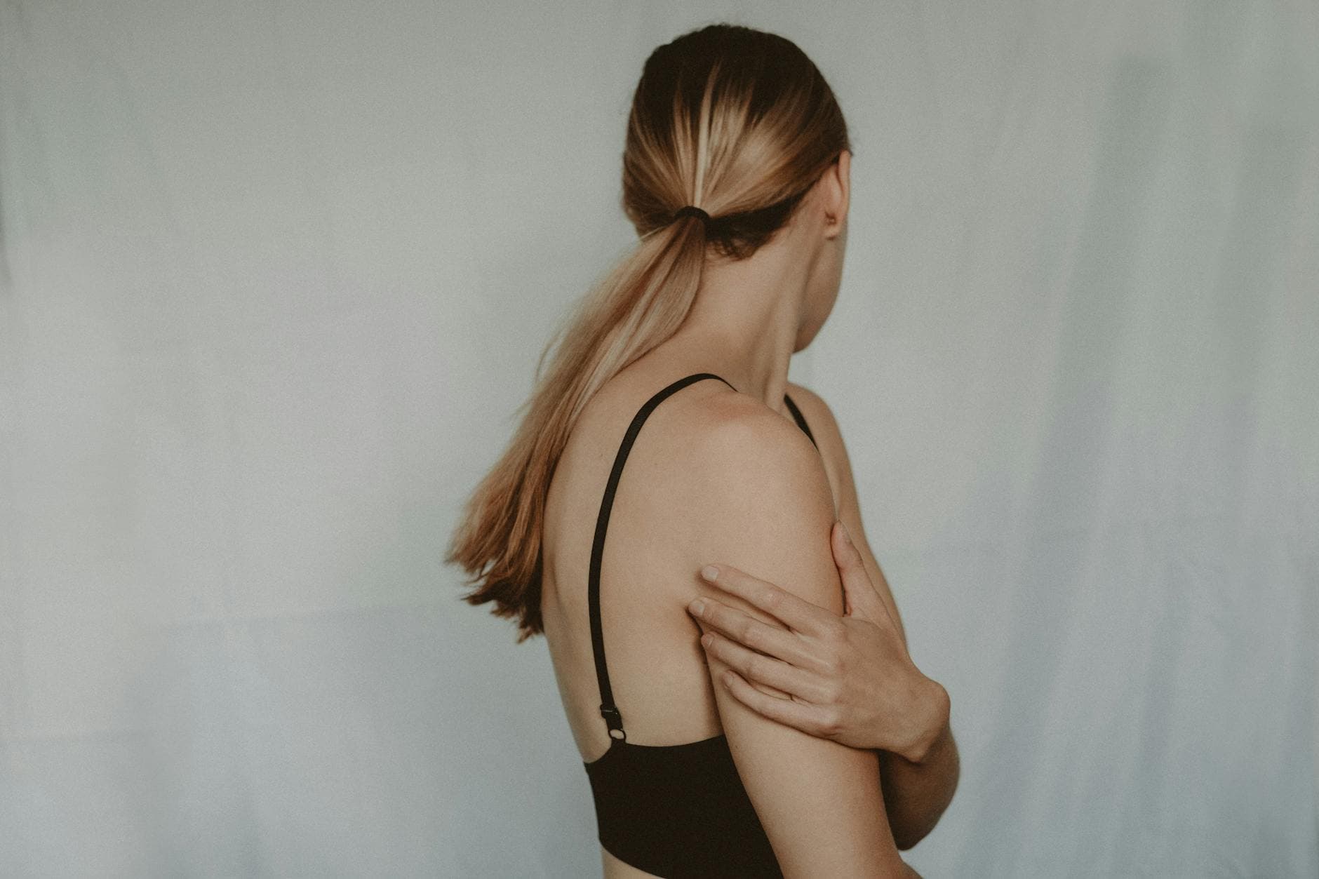 Side view of unrecognizable young female with long hair and black bra standing near grey wall turning head away - therapy for winter apathy