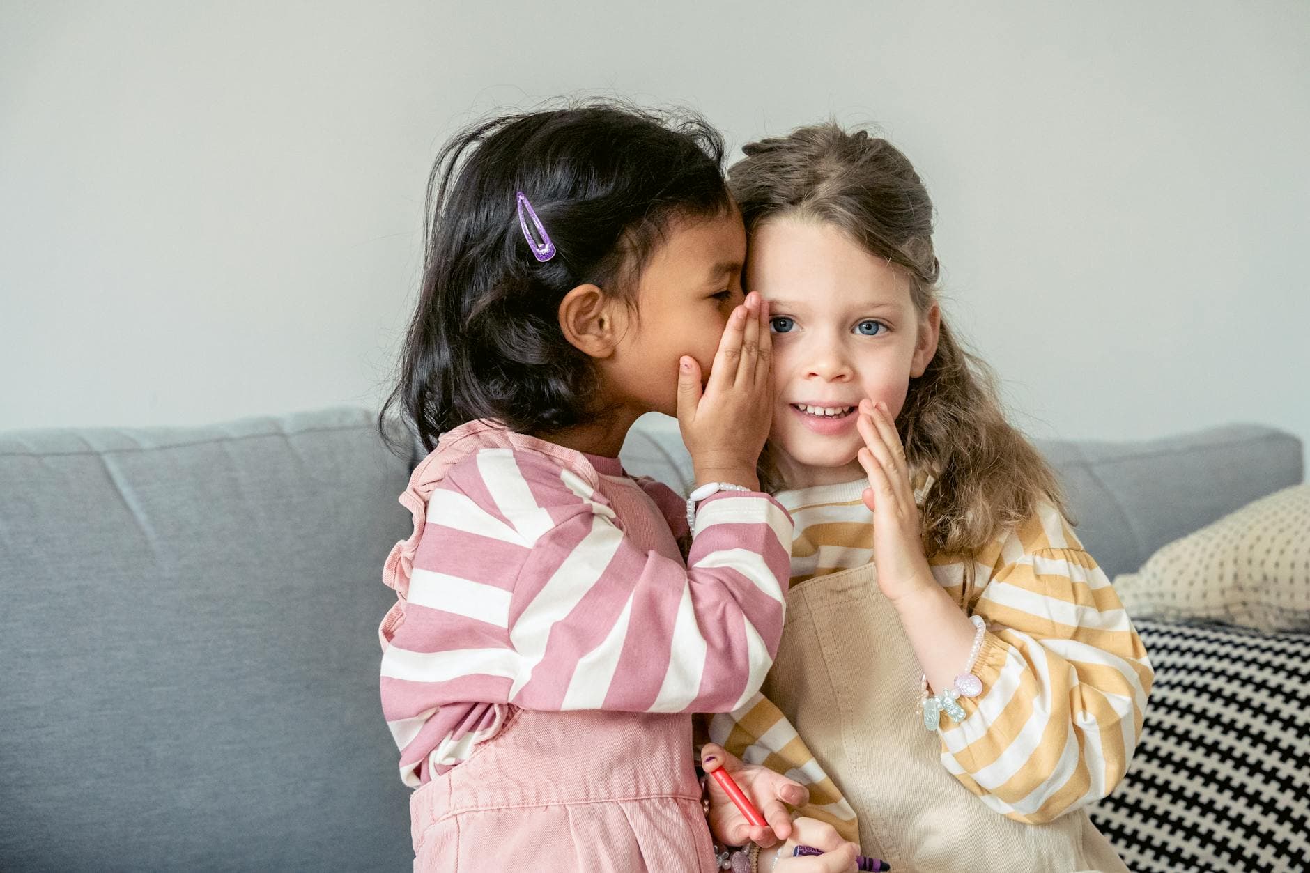 Ethnic girl sitting on sofa close to smiling best friend and whispering secrets on ear - active listening relationship