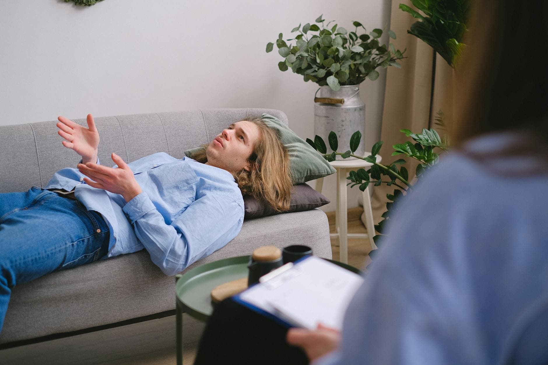 Concerned male client lying on sofa and speaking about mental problems with unrecognizable psychologist with clipboard during psychotherapy appointment in office - cbt for spring procrastination