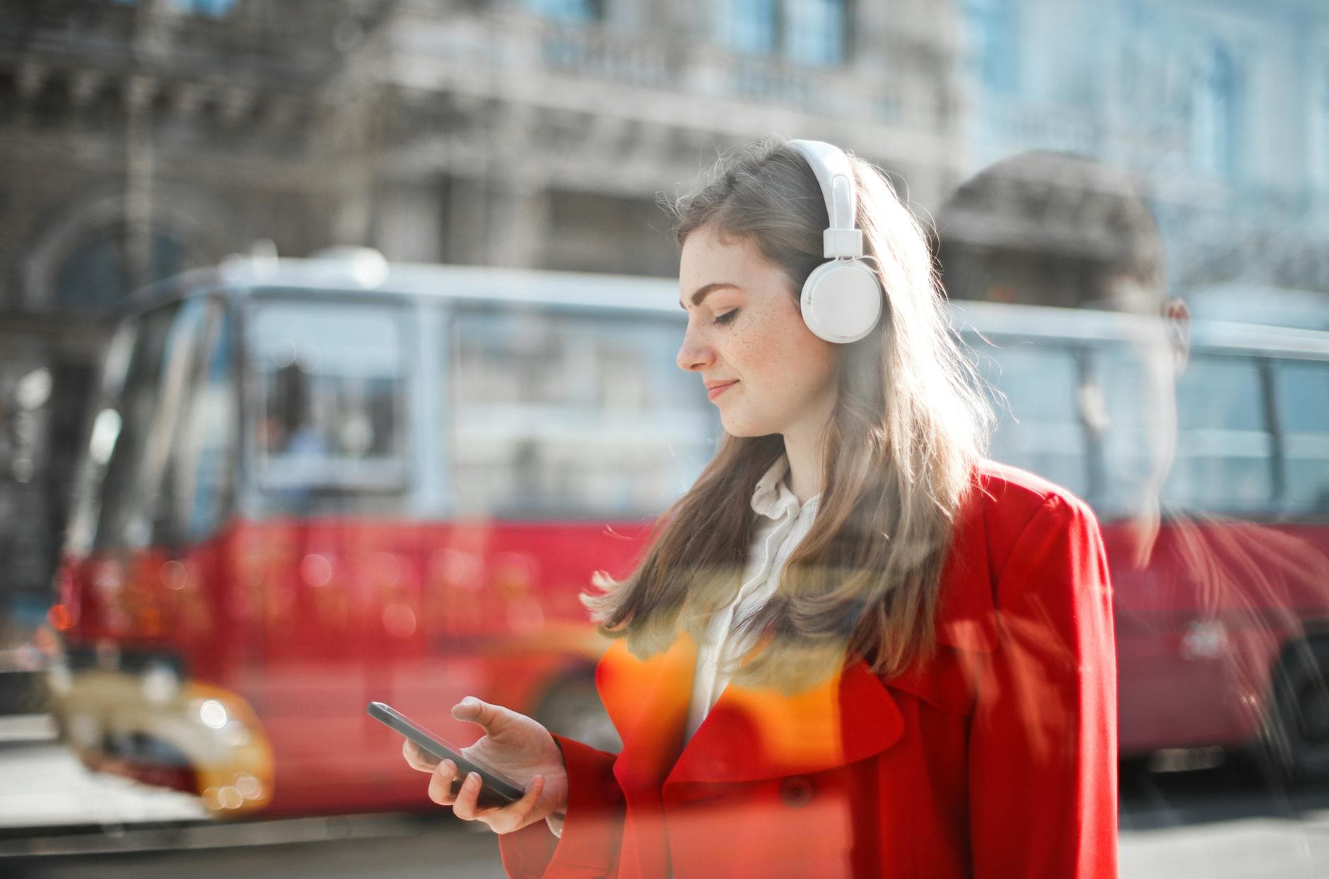 Young woman in red blazer using smartphone and headphones outdoors with bus in background. - effective listening skills