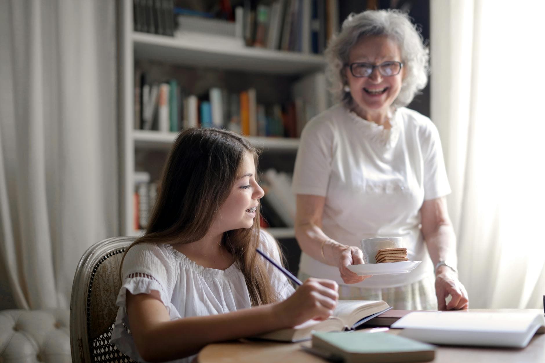 Cheerful grandmother in glasses and casual clothes smiling at camera while giving pastry on plate and mug of tasty beverage to joyful teenager sitting at table with books and exercise book and study in light cozy living room - family dynamics therapy