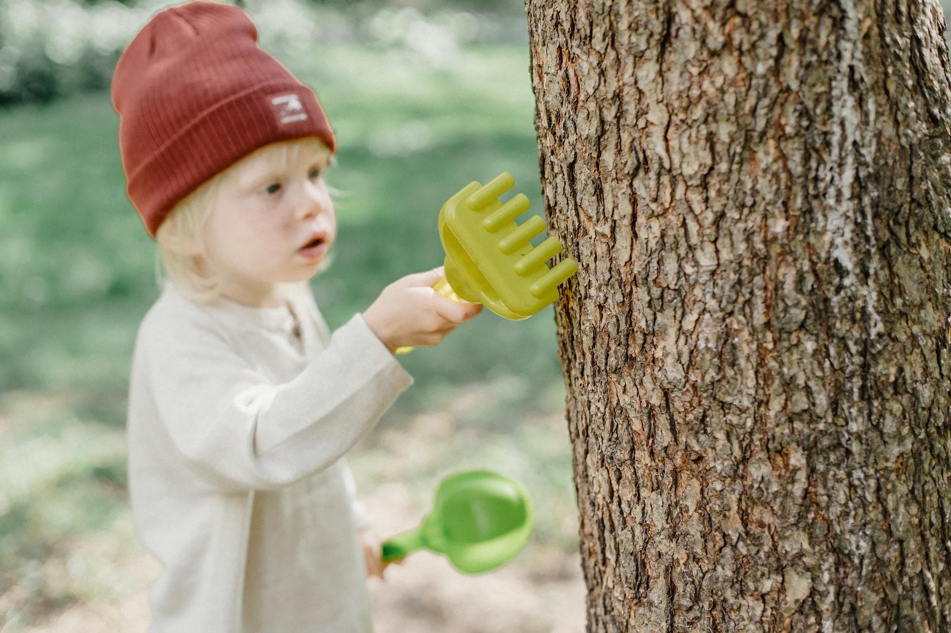 High angle of focused little blond boy in casual wear and brown hat rubbing tree trunk with toy rake in garden - toddler outdoor activities
