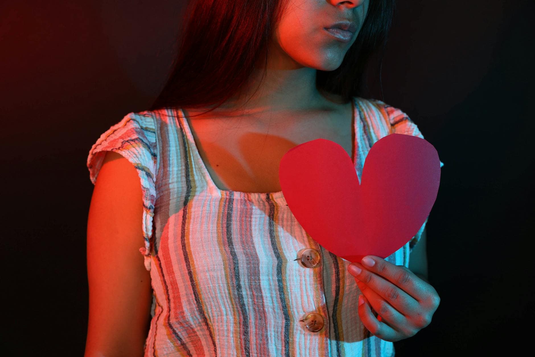 Young woman holding a red paper heart, portrayed in dramatic red and blue lighting. - post breakup healing