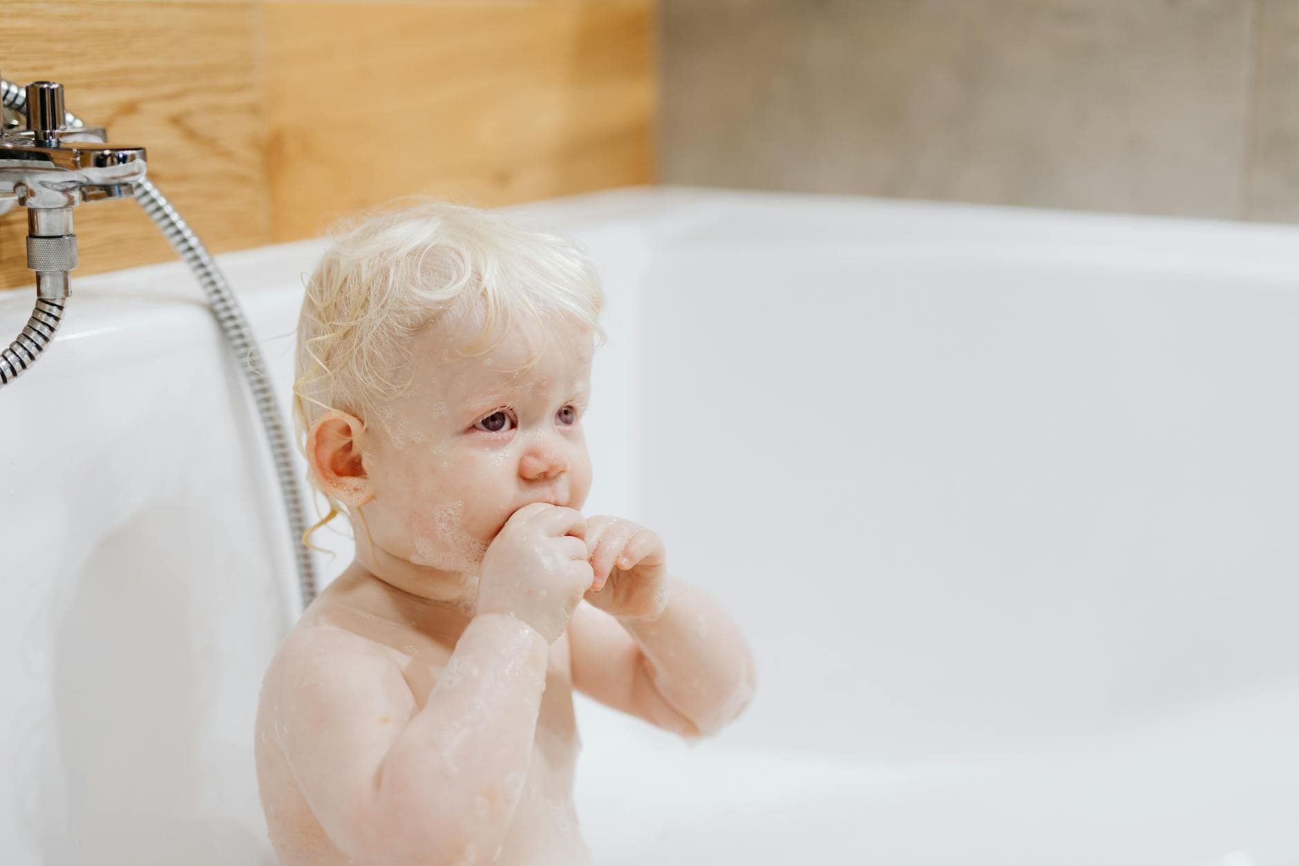 Cute baby with blonde hair enjoying a bath in a white bathtub indoors. - toddler self care