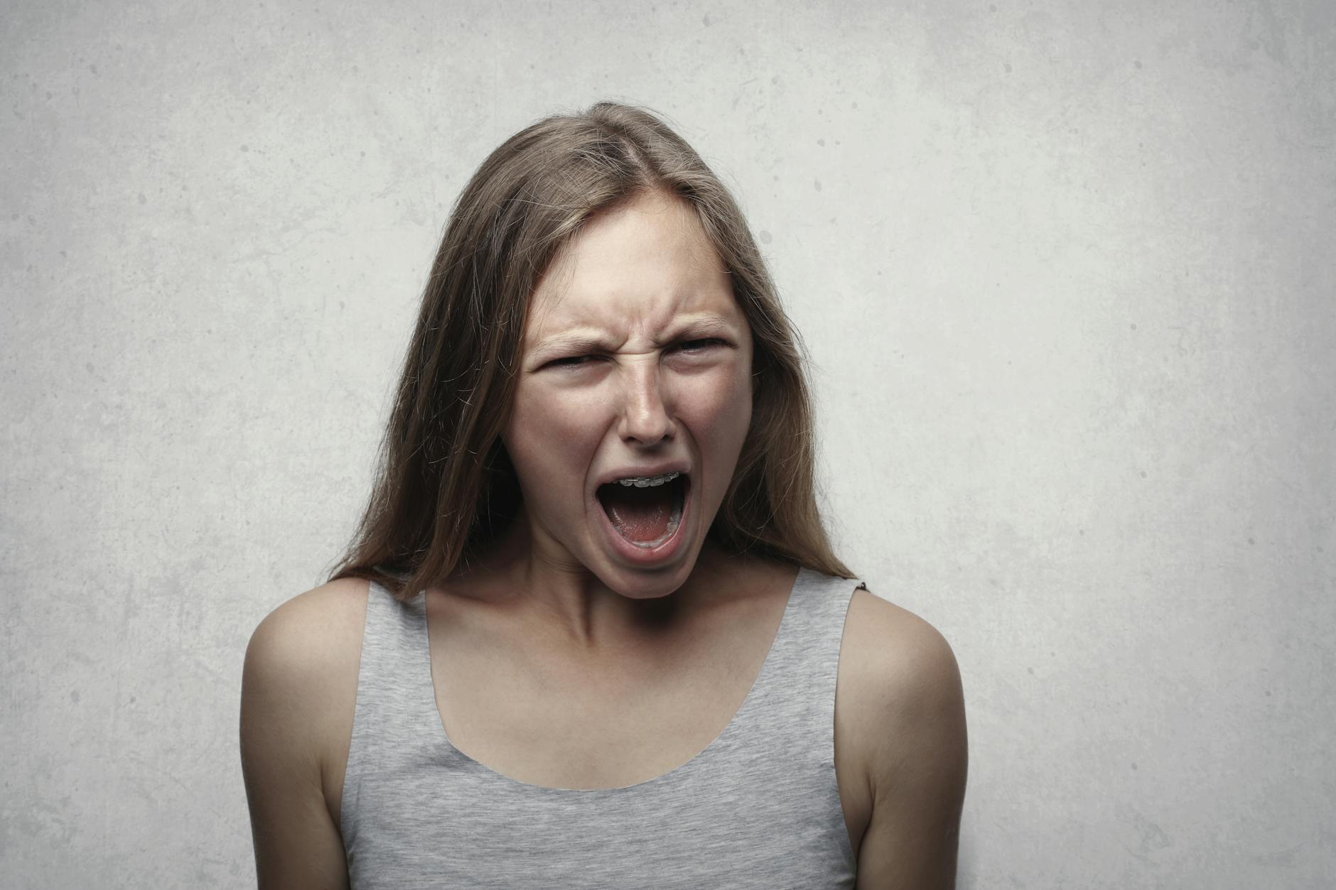 A woman with an intense facial expression screaming against a neutral gray background. - anger management near me