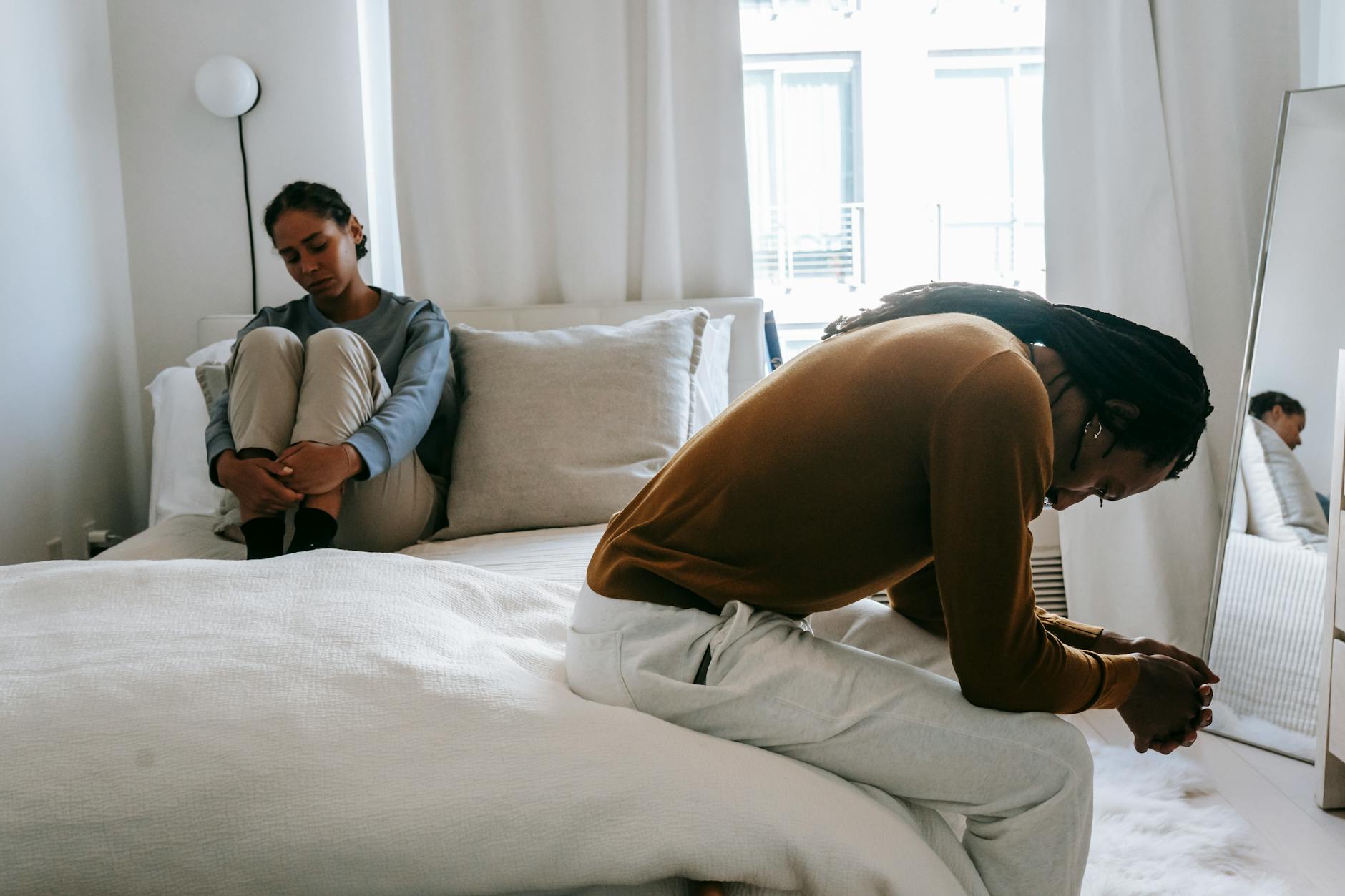 Discontented black man with clasped hands sitting on bed near upset girlfriend during quarrel in apartment - anxiety in romantic relationships