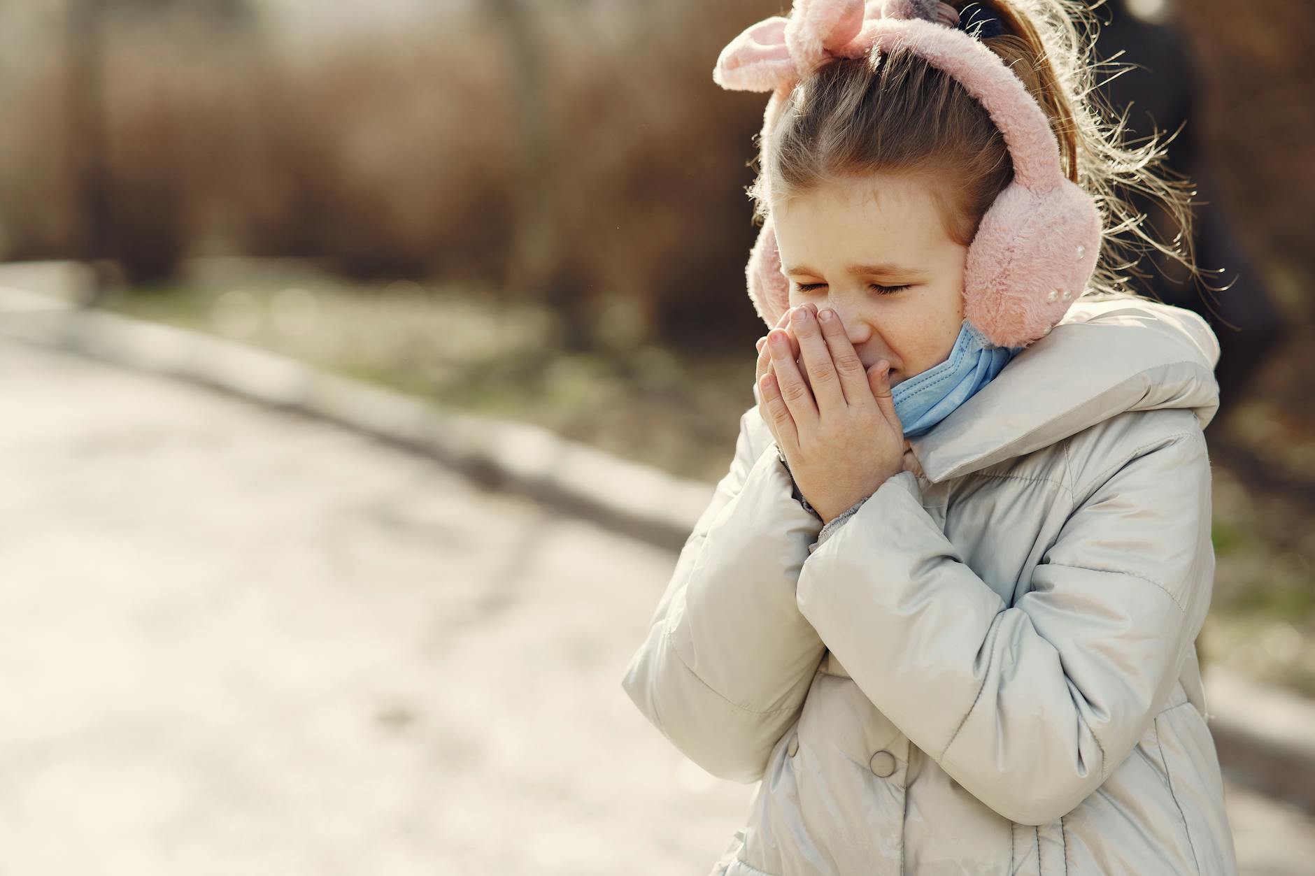 Small child with lowered face mask covering face by hands while sneezing on street - childhood trauma symptoms