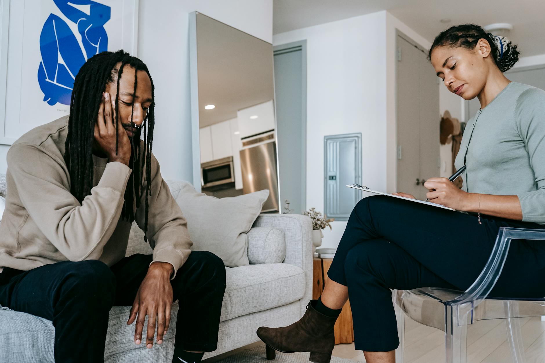 Attentive African American female with clipboard taking notes near sad male partner touching face on couch at home - choose therapist seasonal depression