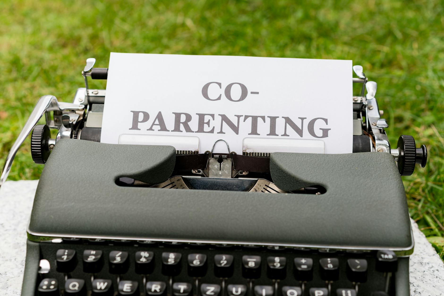 Close-up of an antique typewriter with 'Co-Parenting' paper, set outdoors on grass. - co-parenting communication winter break