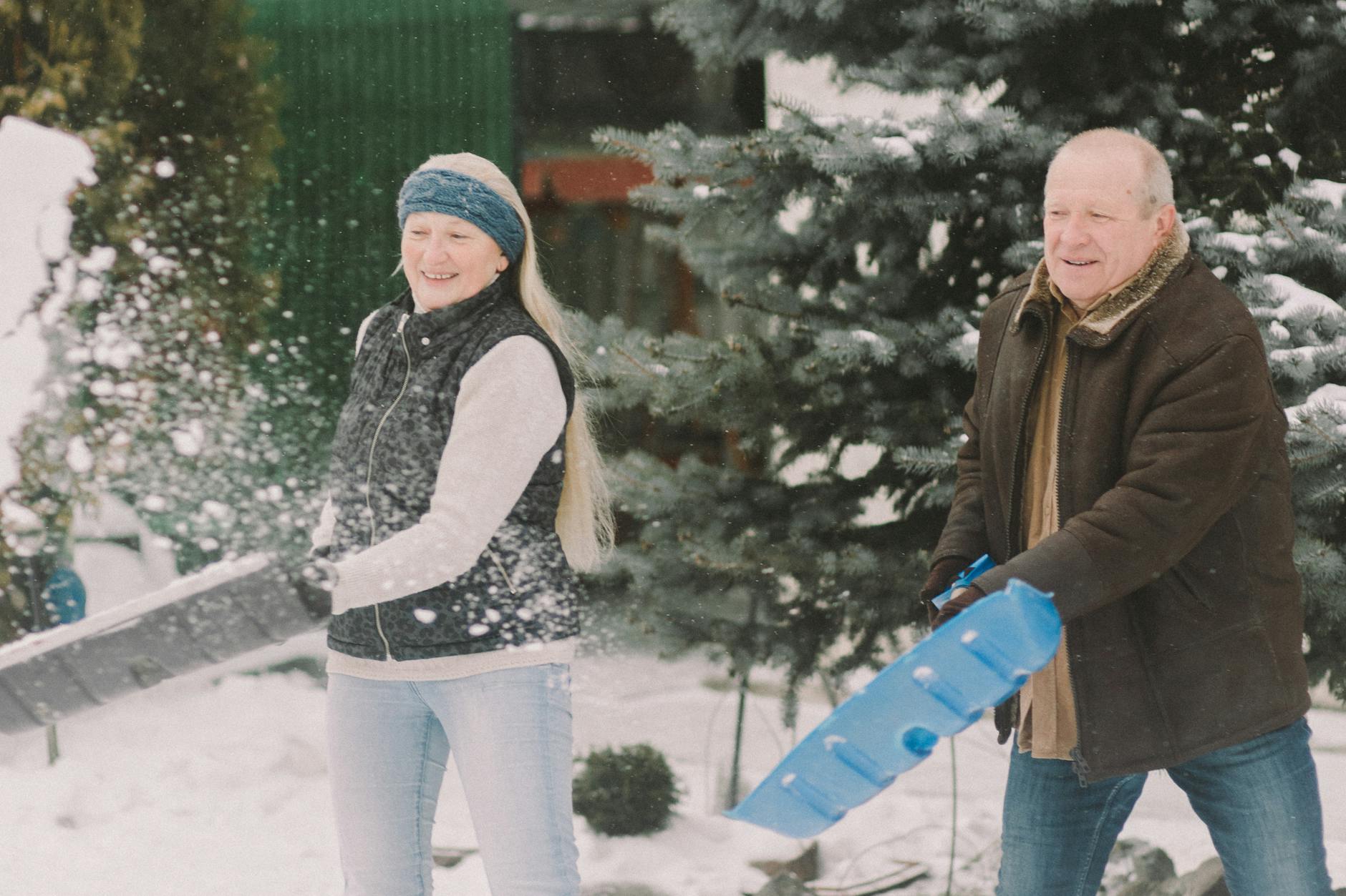 An elderly couple happily shovels snow together outdoors, embracing the winter season. - cold weather couple communication