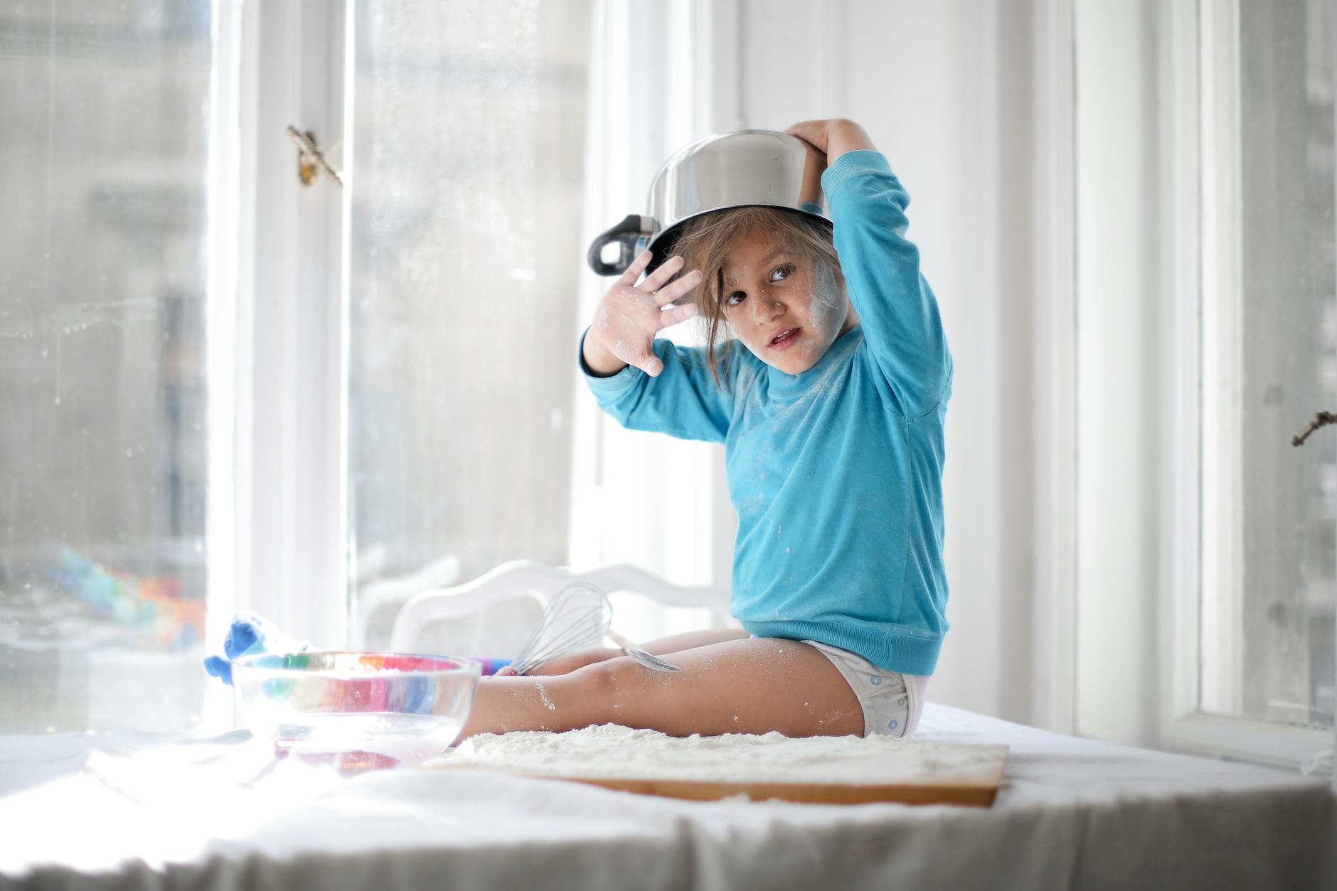 A child in a blue sweater playfully wears a pot on their head while sitting on a kitchen table. - creative indoor play snowy days