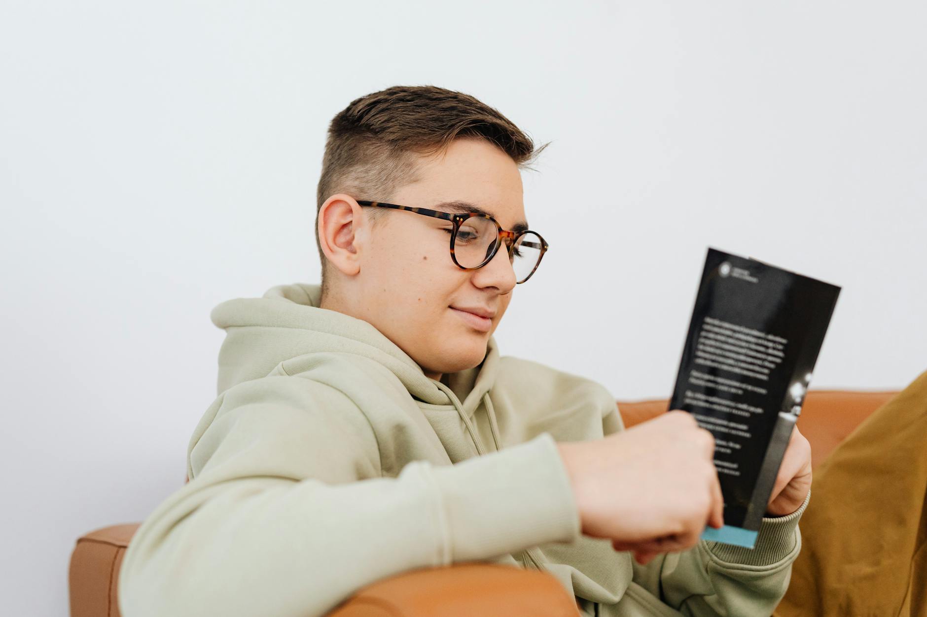 A young man wearing glasses enjoys reading a book while relaxing indoors. - improving teen communication at home