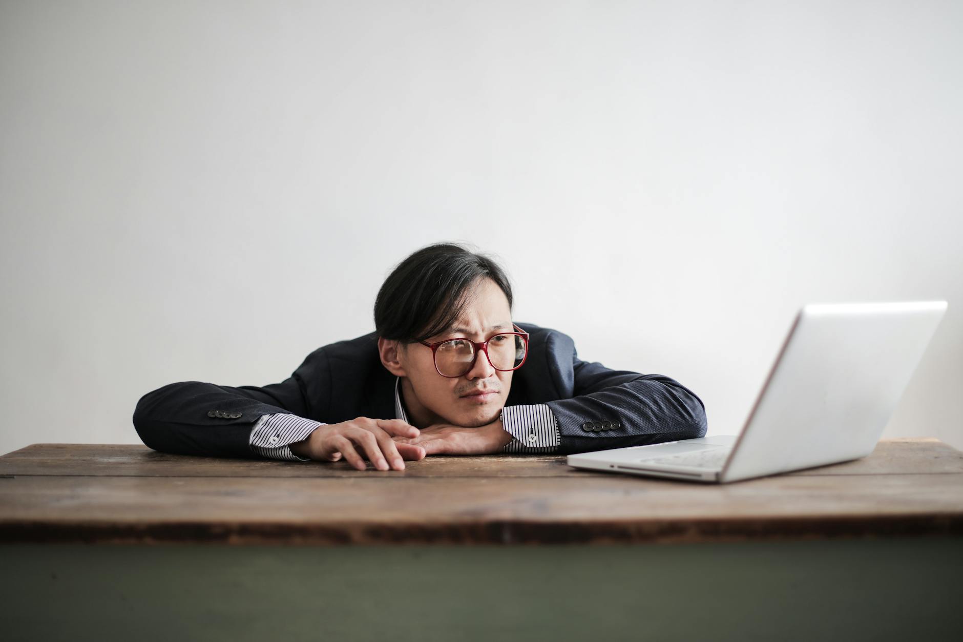 Asian businessman in glasses, resting head on desk, looking at laptop in office setting. - kids winter break boredom