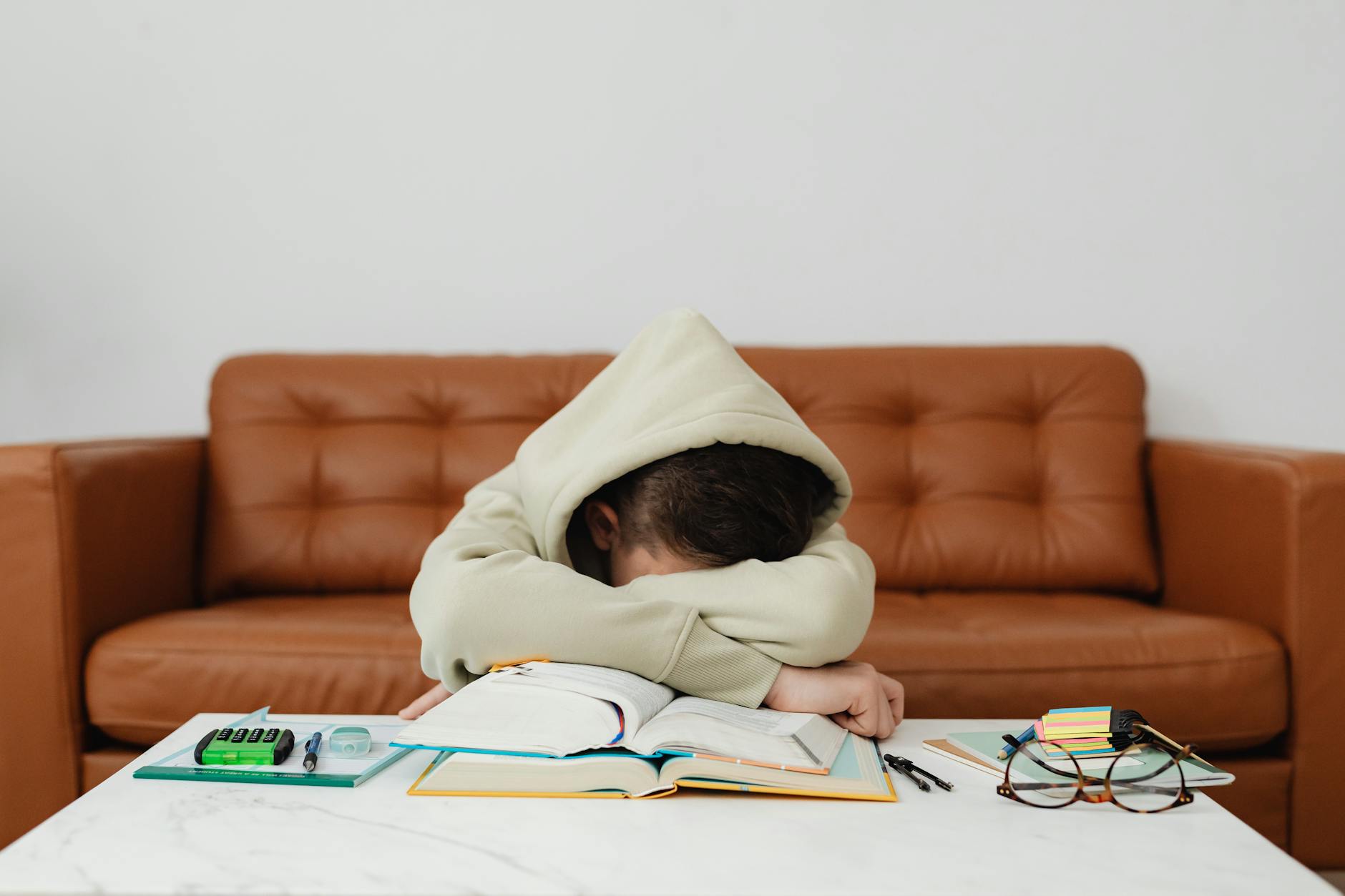 Child in hoodie leans over books, exhausted while studying on a couch. - manage kids winter frustration