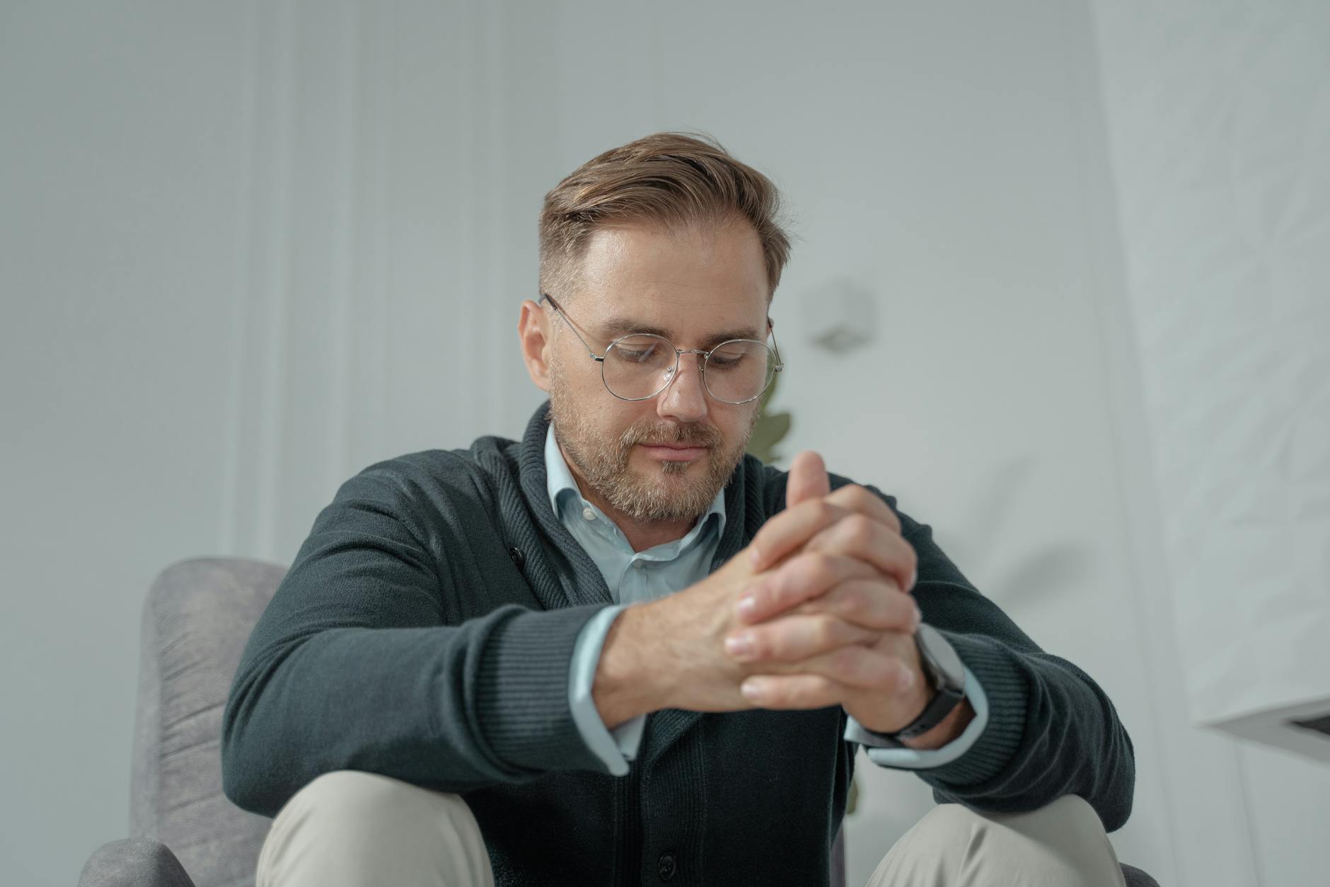 A thoughtful man with glasses and gray sweater sitting indoors, hands clasped. - mindfulness for winter anxiety