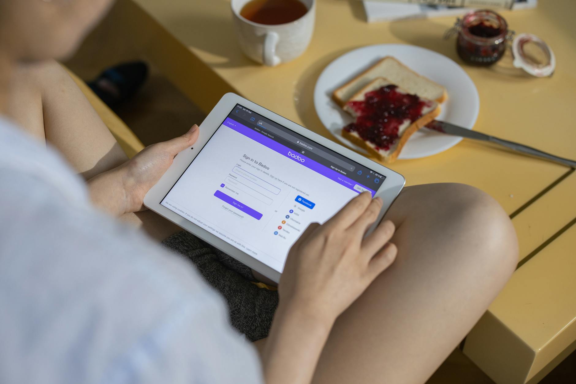 A woman using a tablet while having a snack with jam on toast and tea, reflecting a cozy indoor setting. - online dating tips men