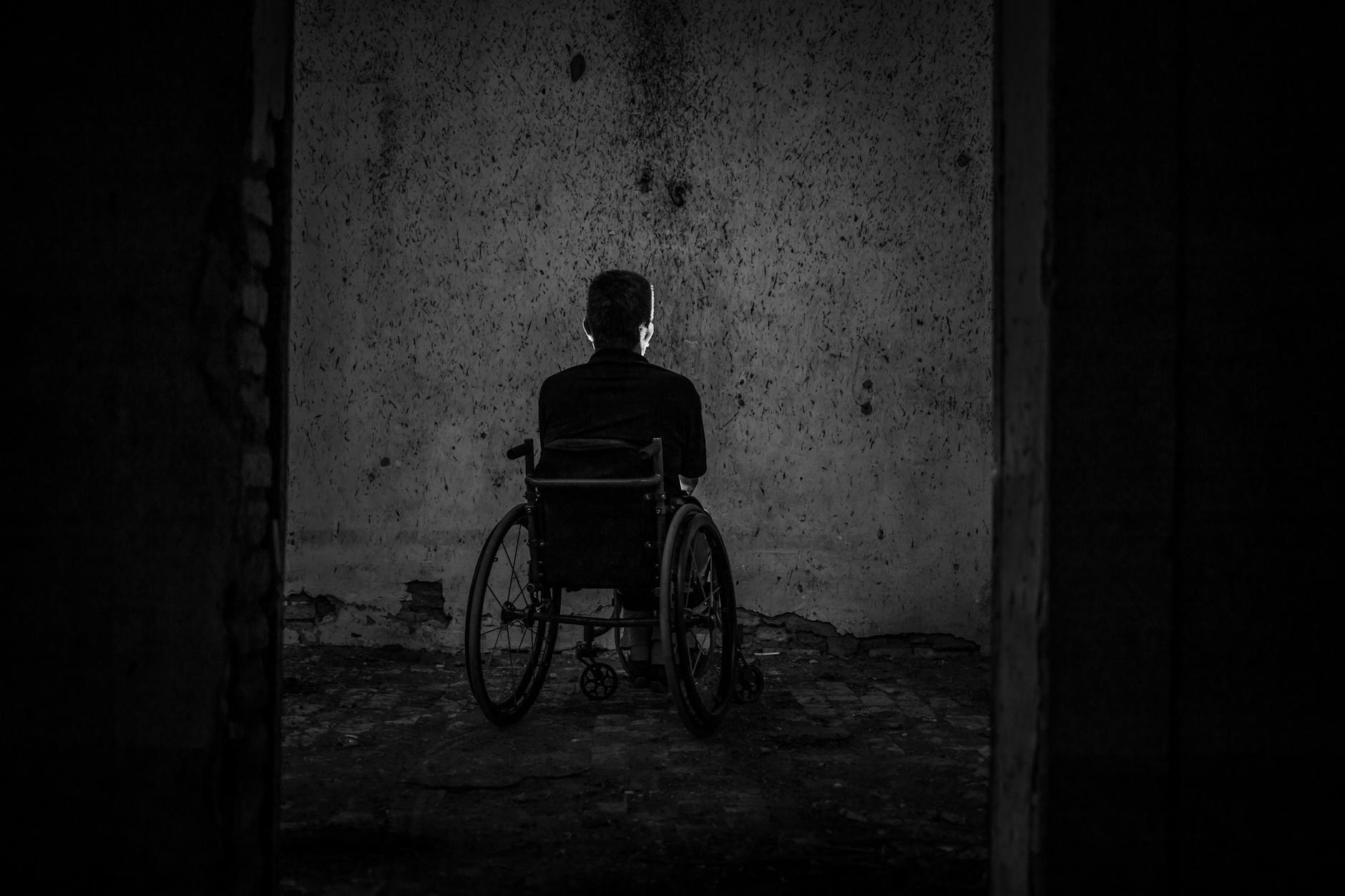 A man sits alone in a wheelchair facing a wall in a dark, empty room, evoking solitude. - overcome winter isolation