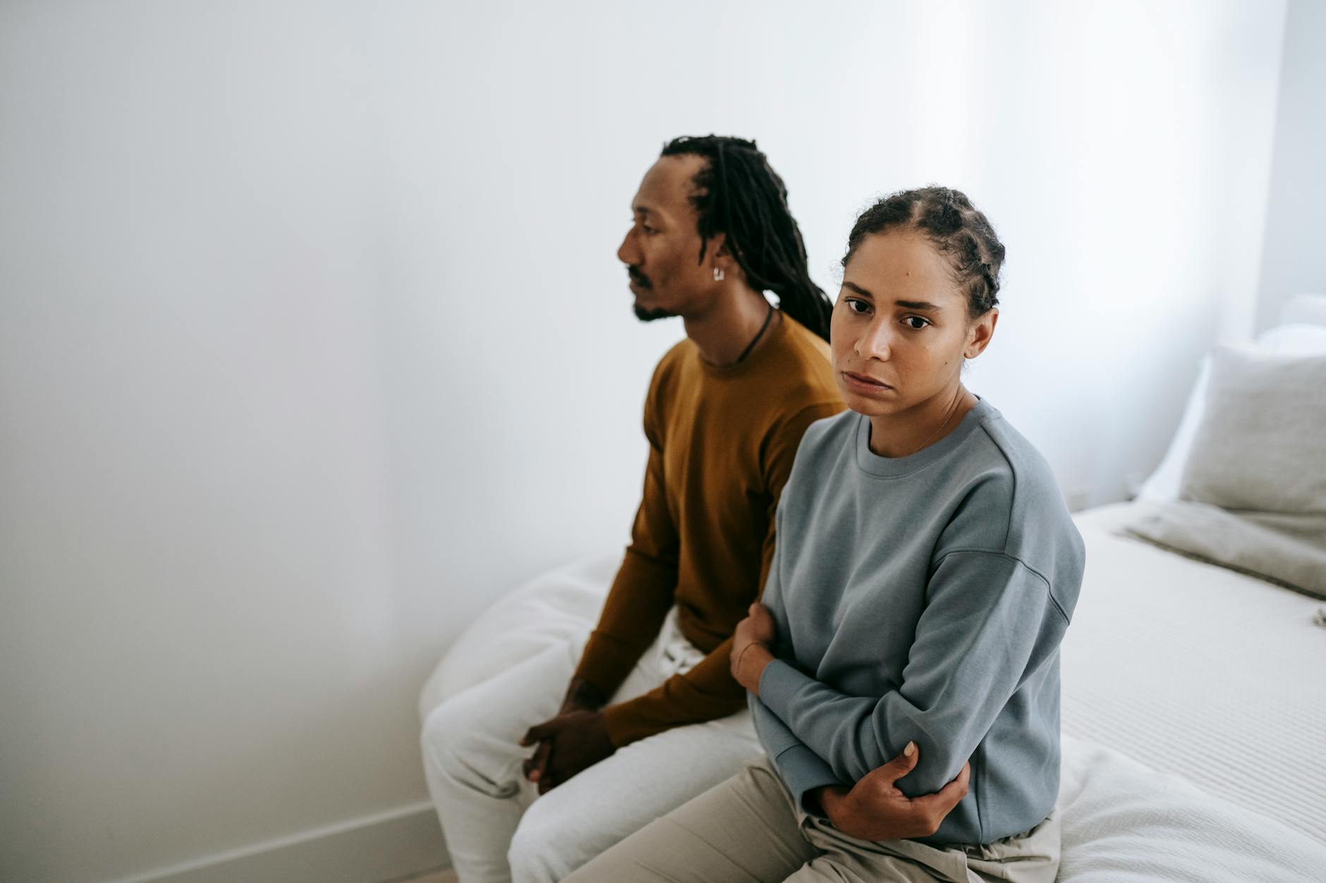 A couple sitting silently on a bed with tense expressions, suggesting conflict and emotional struggles. - post-holiday relationship struggles