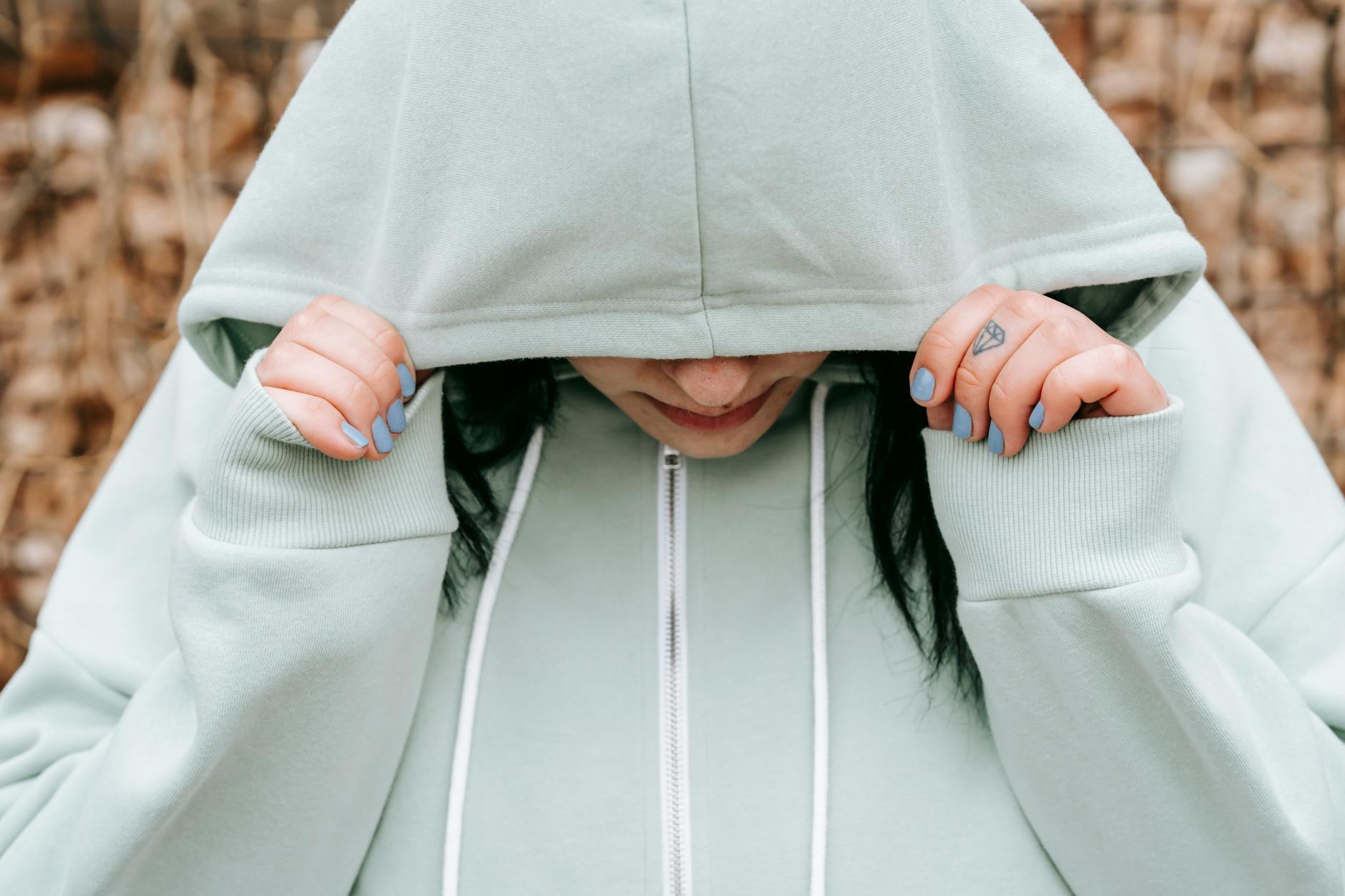 Crop unhappy female covering lowered head with hood in autumn forest in daylight - seasonal depression teens