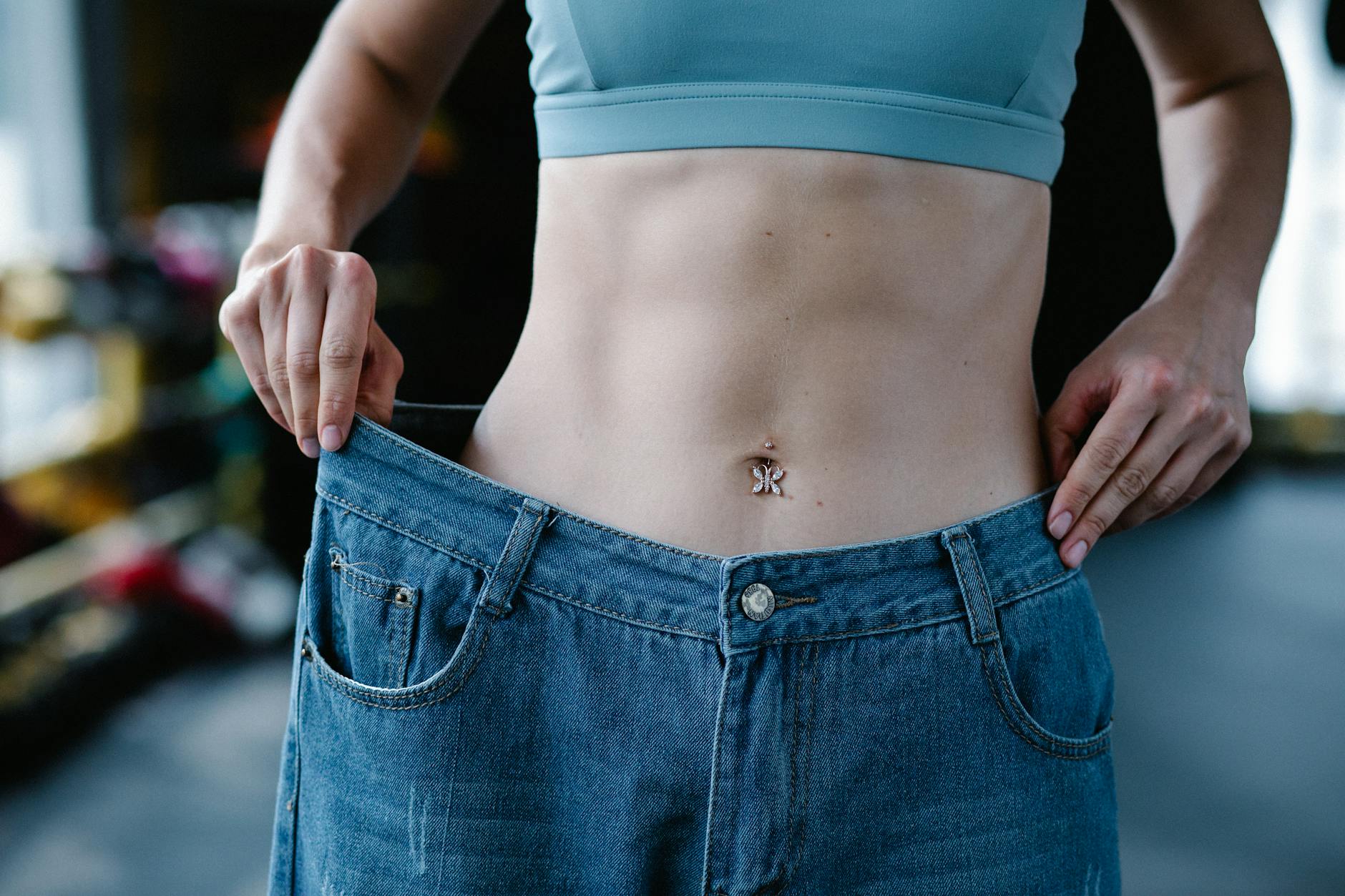 A woman shows her weight loss by holding oversized jeans revealing her toned stomach. - self esteem building exercises
