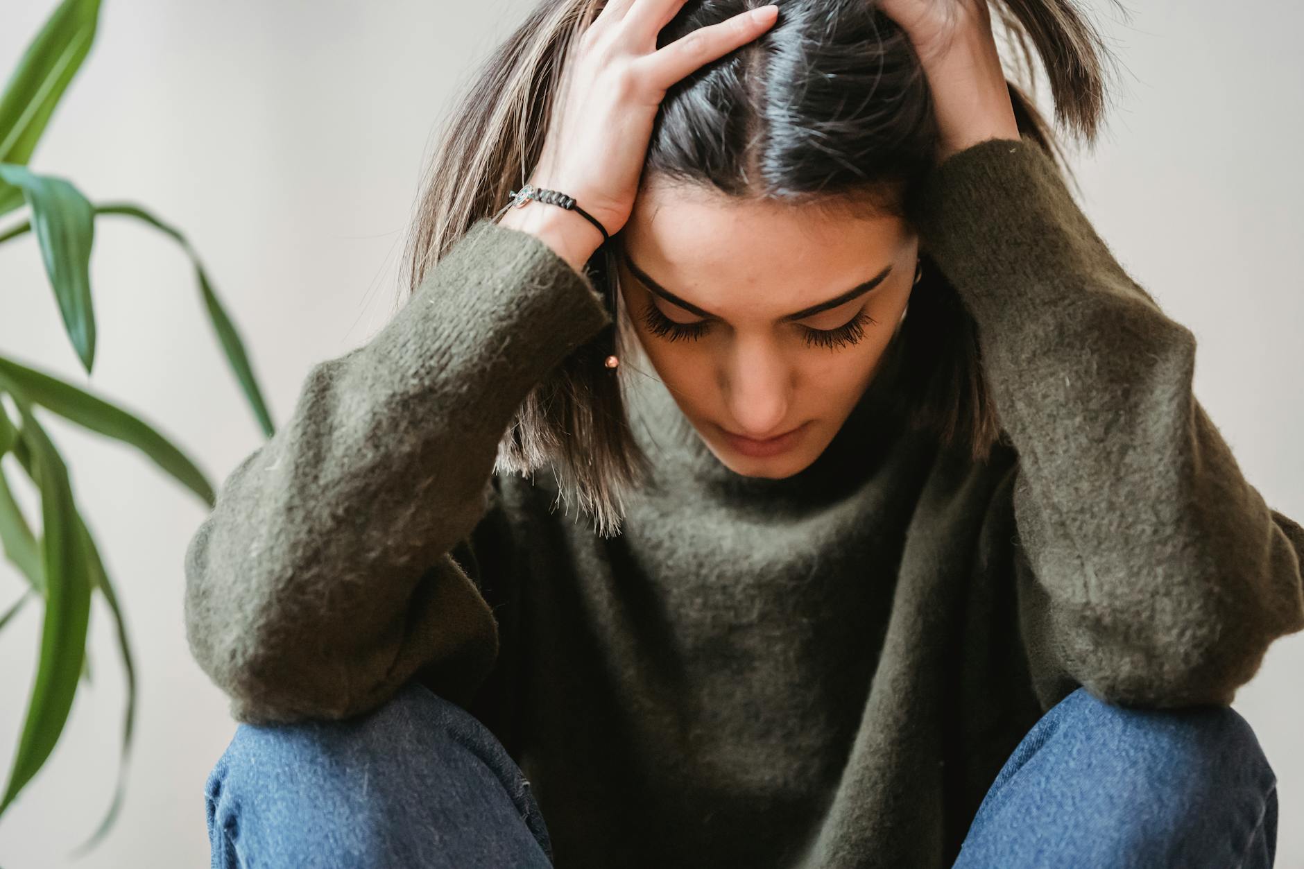 Crop disappointed young ethnic female with dark hair in casual clothes grabbing head with hands and looking down while sitting against white wall at home - therapy for winter apathy