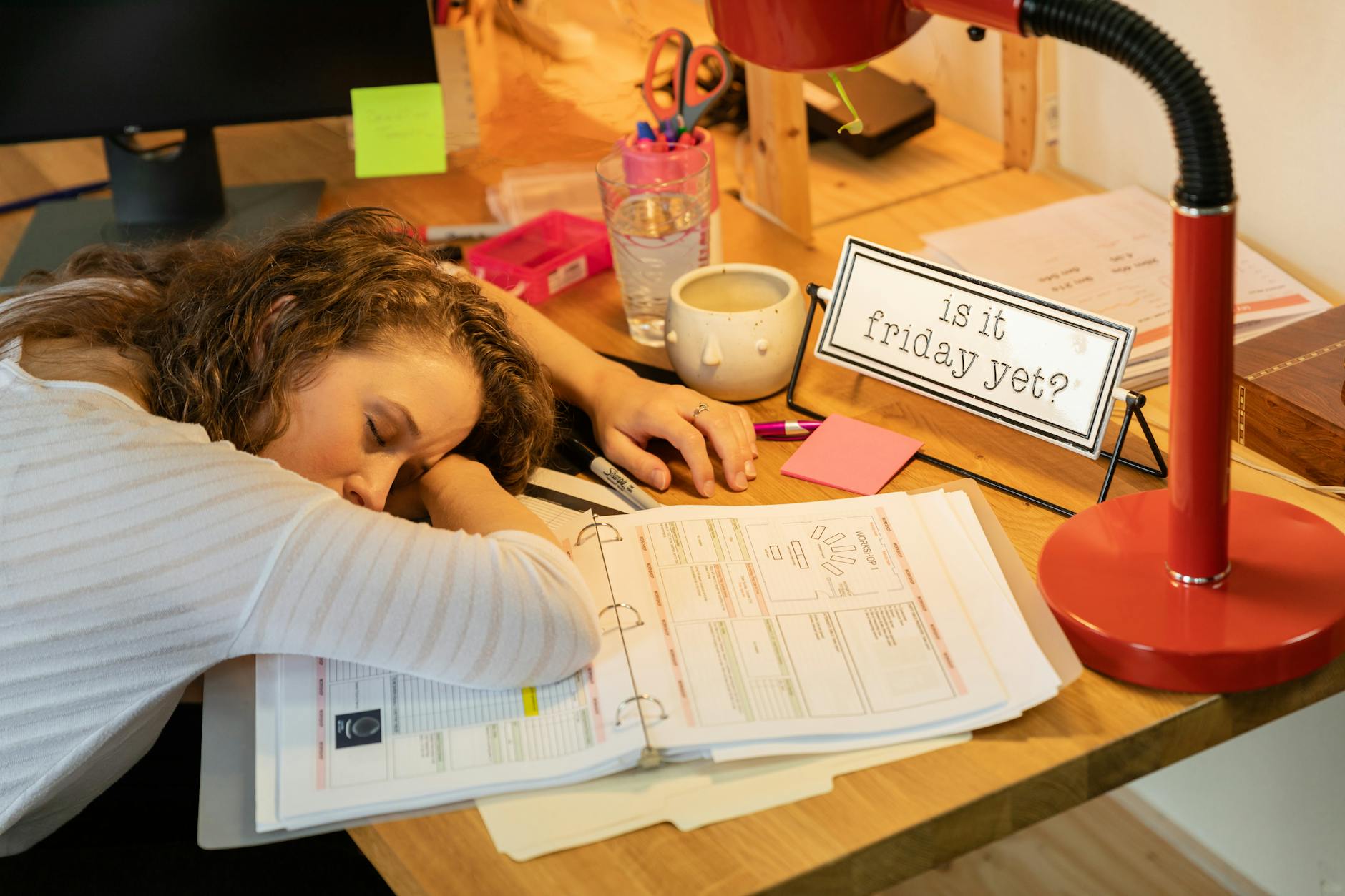 A woman sleeping at her desk with office documents, a coffee cup, and a red desk lamp. - therapy for winter slump