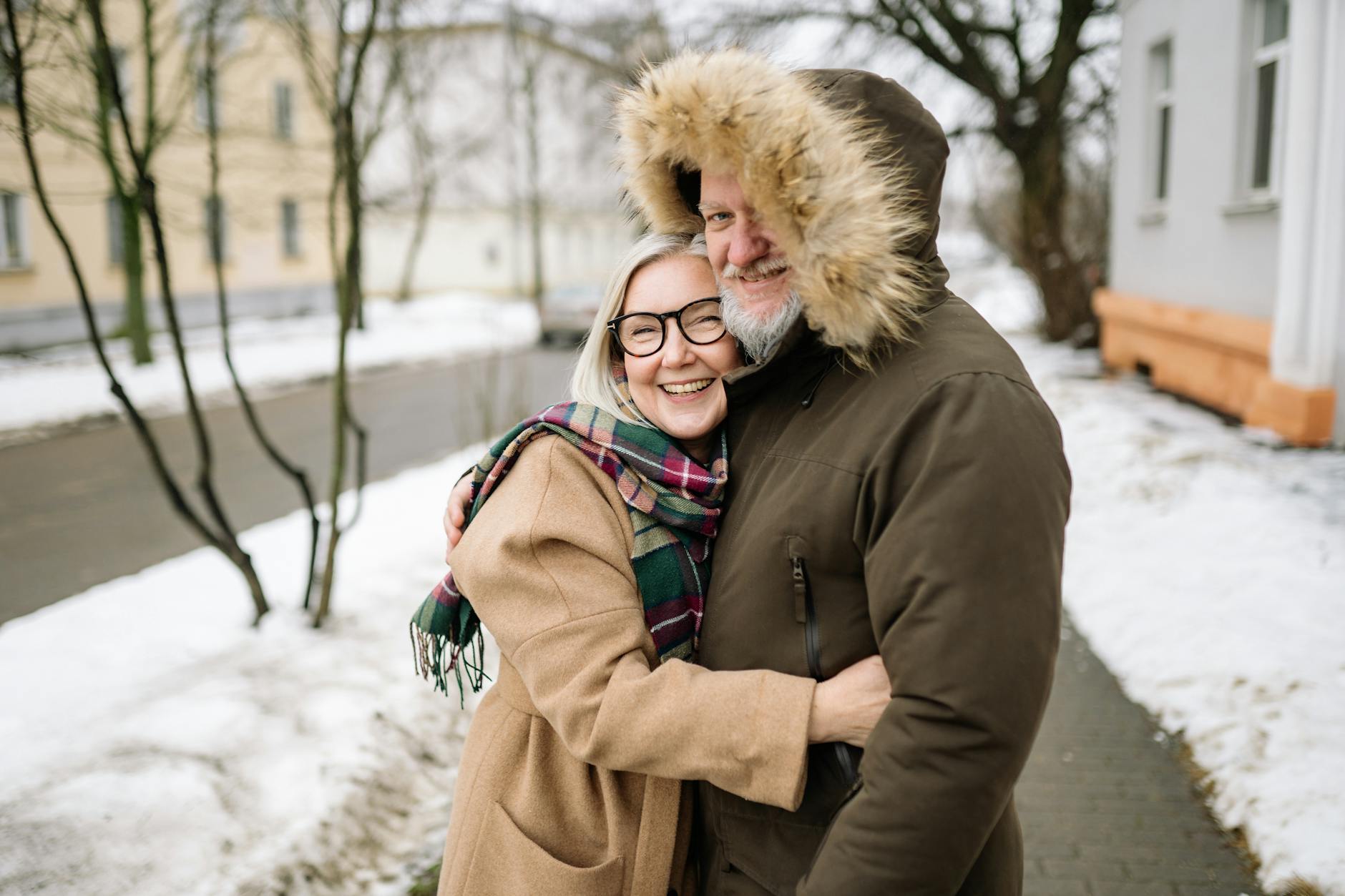 Happy senior couple in winter coats smiling and embracing outdoors on a snowy day. - winter happiness couples