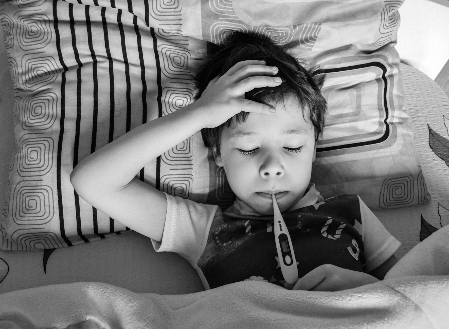 Black and white overhead view of child in casual clothing measuring body temperature with electronic thermometer while lying on bed under blanket - winter sleep problems