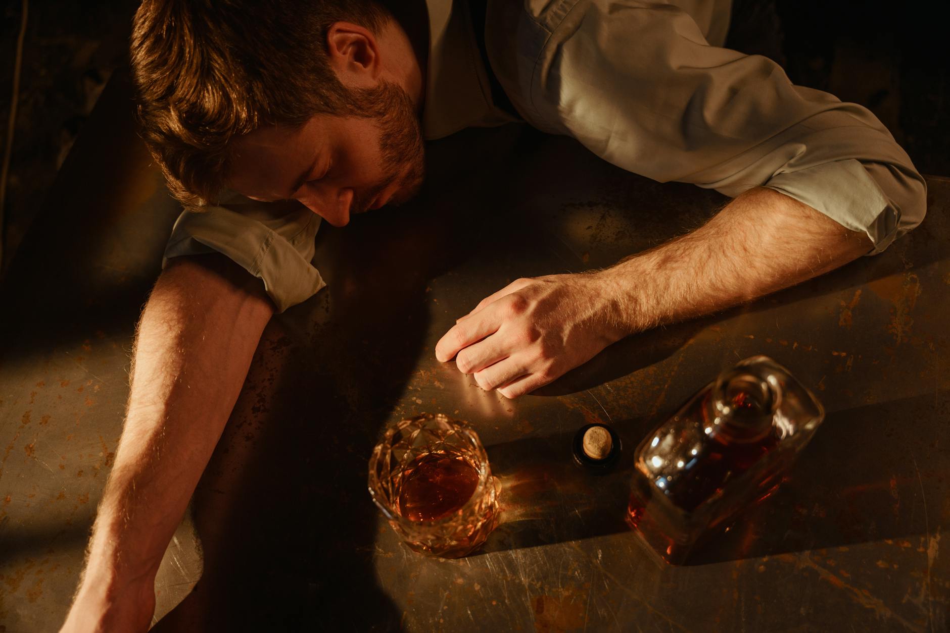 A man sleeps on a table with a whiskey glass and bottle nearby, suggesting exhaustion. - winter sleep problems