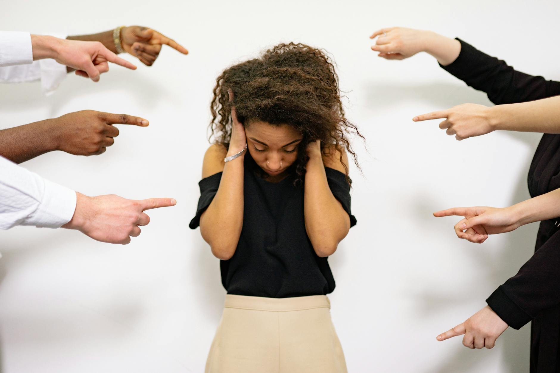 A young woman with curly hair looks distressed as multiple hands point accusatory fingers at her. - winter social anxiety