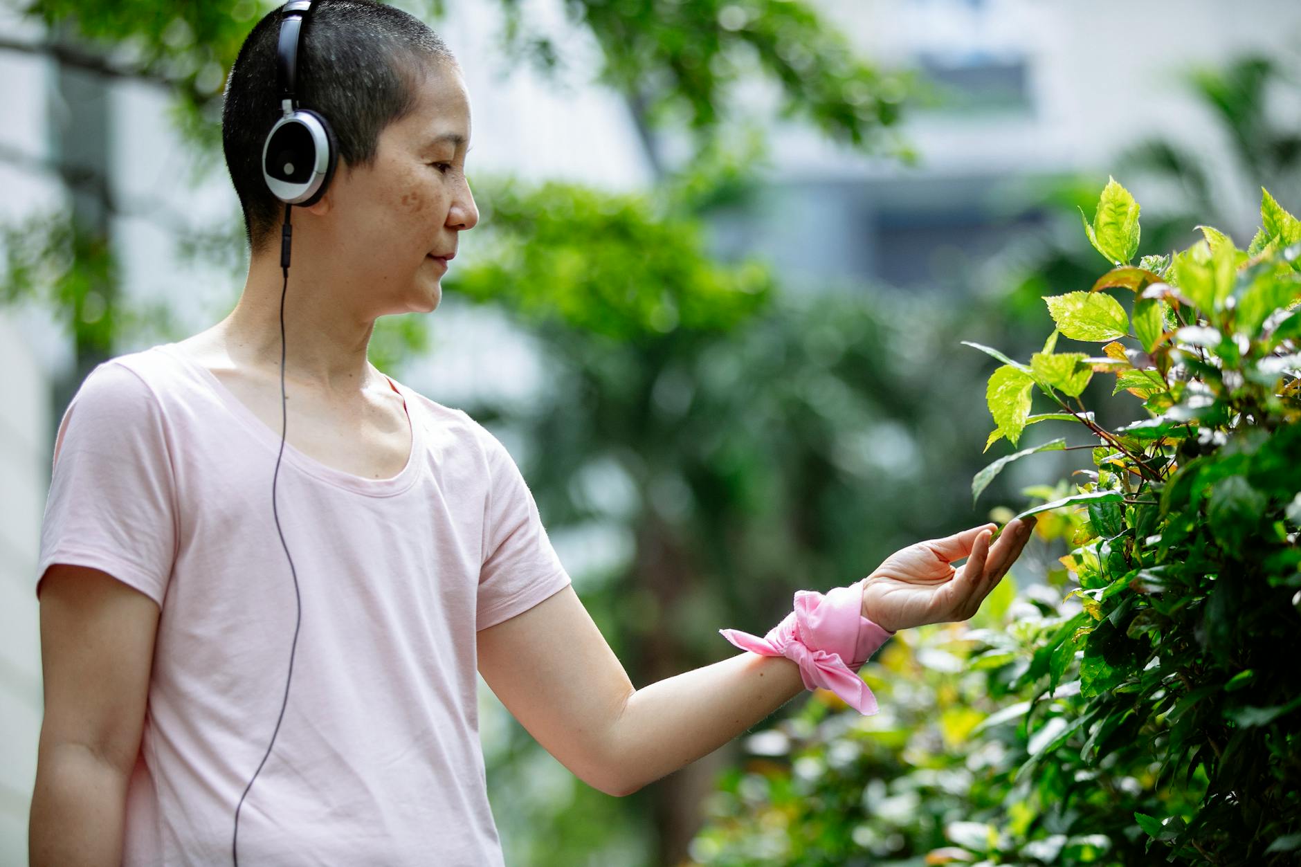Thoughtful middle age ethnic woman in casual clothes with headphones and pink bracelet examining bushes and plants with green leaves in park in summer day - active listening definition