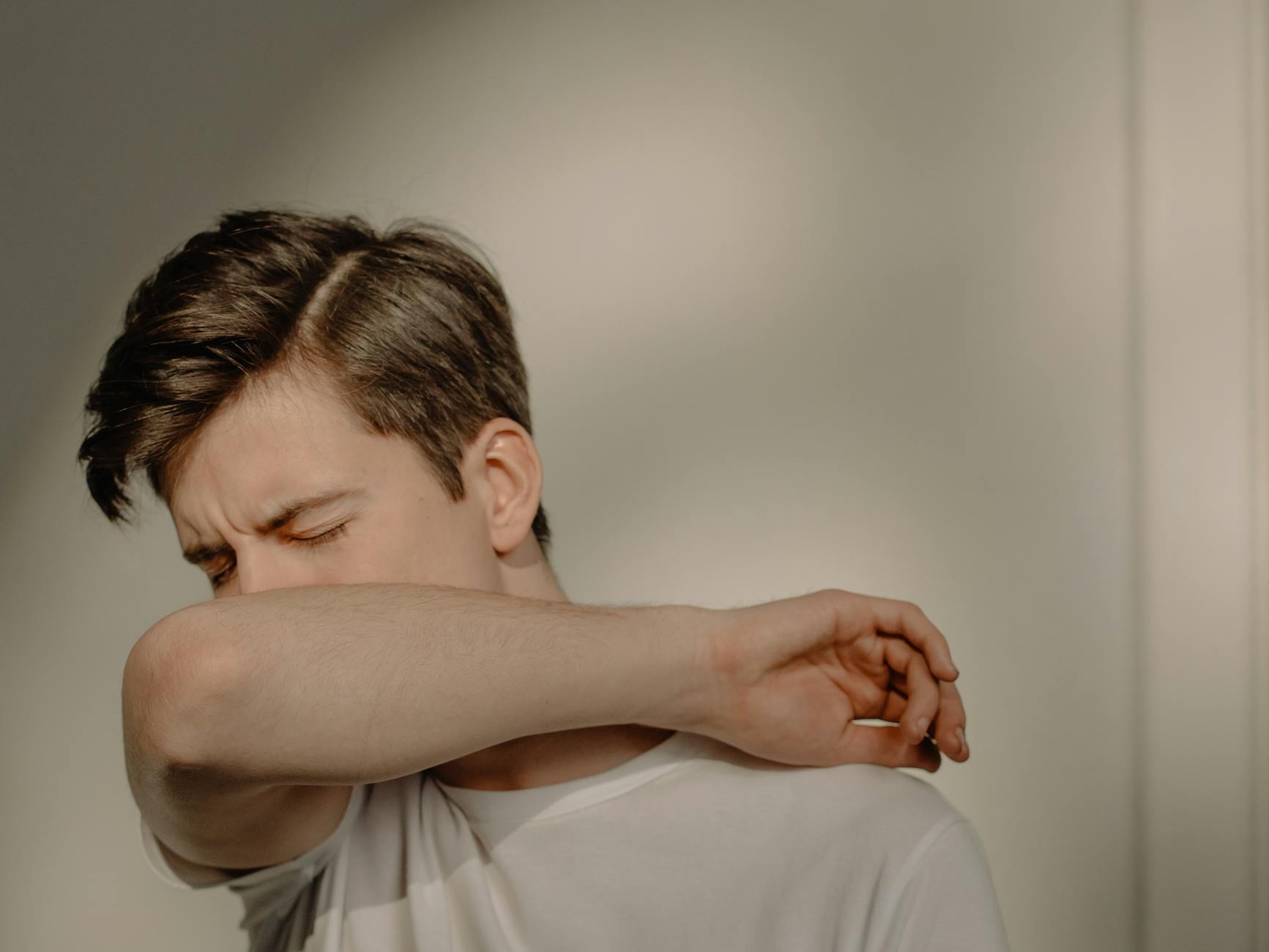 A young man sneezes into his elbow, showcasing a hygiene practice, in an indoor setting. - allergy mood changes