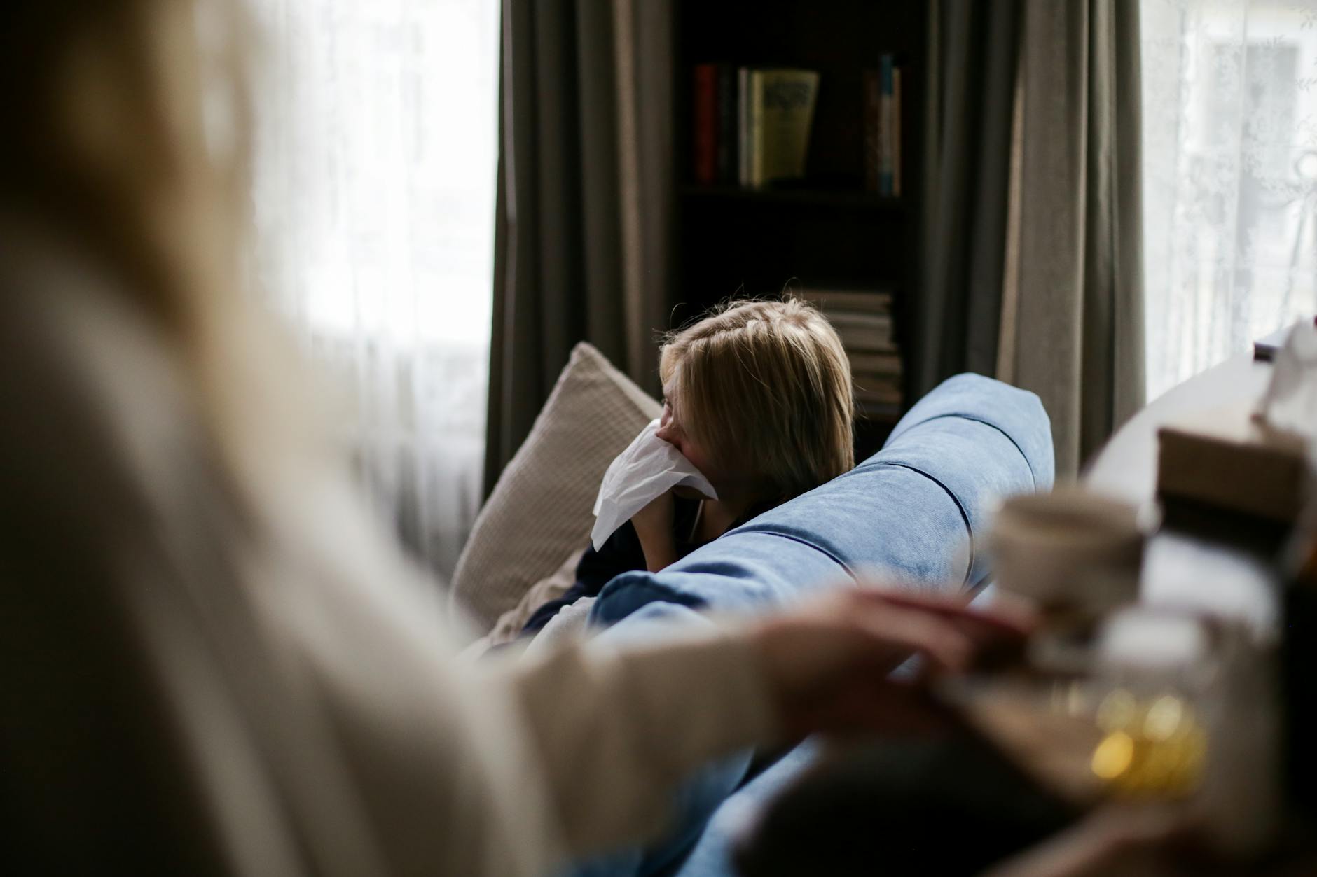 A young child sits on a couch indoors, using a tissue, suggesting illness or allergy. - allergy mood changes