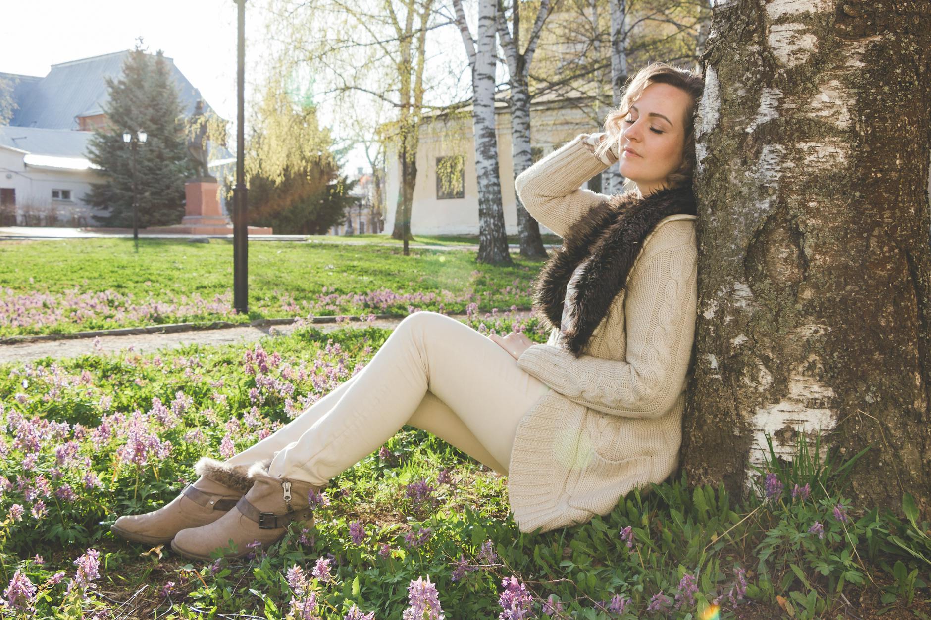 A woman enjoys a peaceful moment sitting against a tree in a spring park, surrounded by blooming flowers. - allergy season relationship
