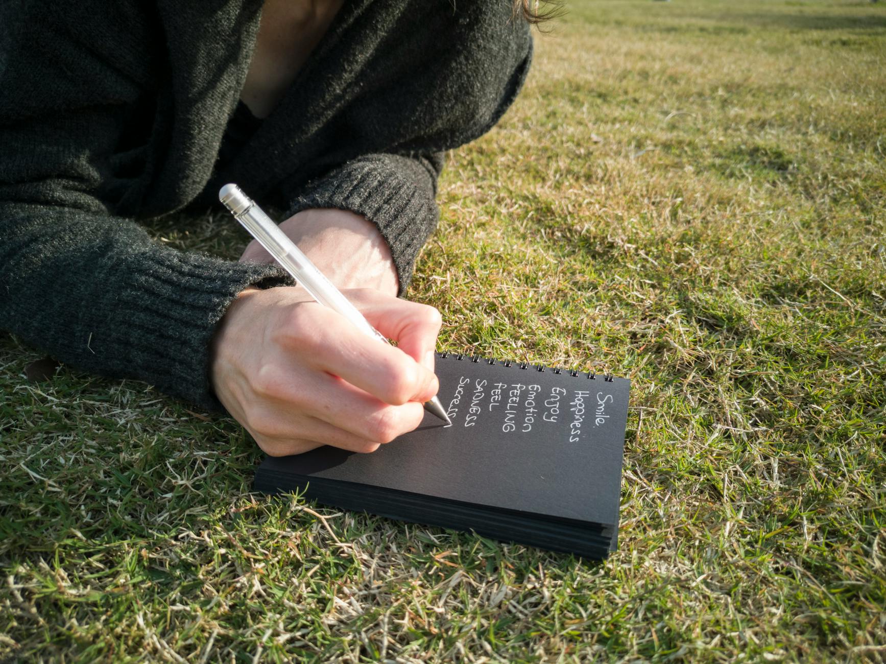 Close-up of a person writing emotions in a notebook while lying on grass outdoors. - cbt for clarity