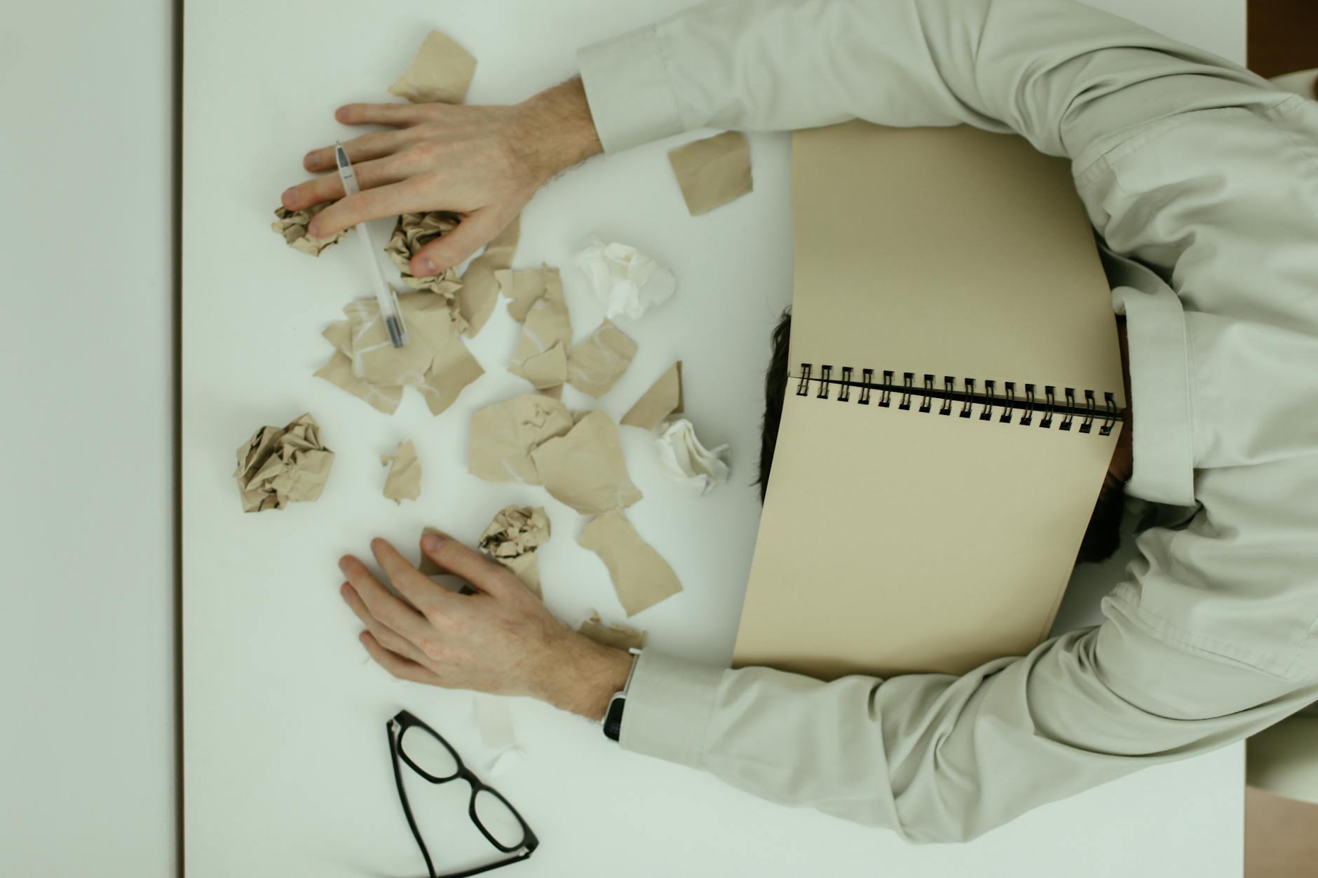 A tired adult man resting his head on a desk cluttered with crumpled papers and an open notebook. - clear mental clutter
