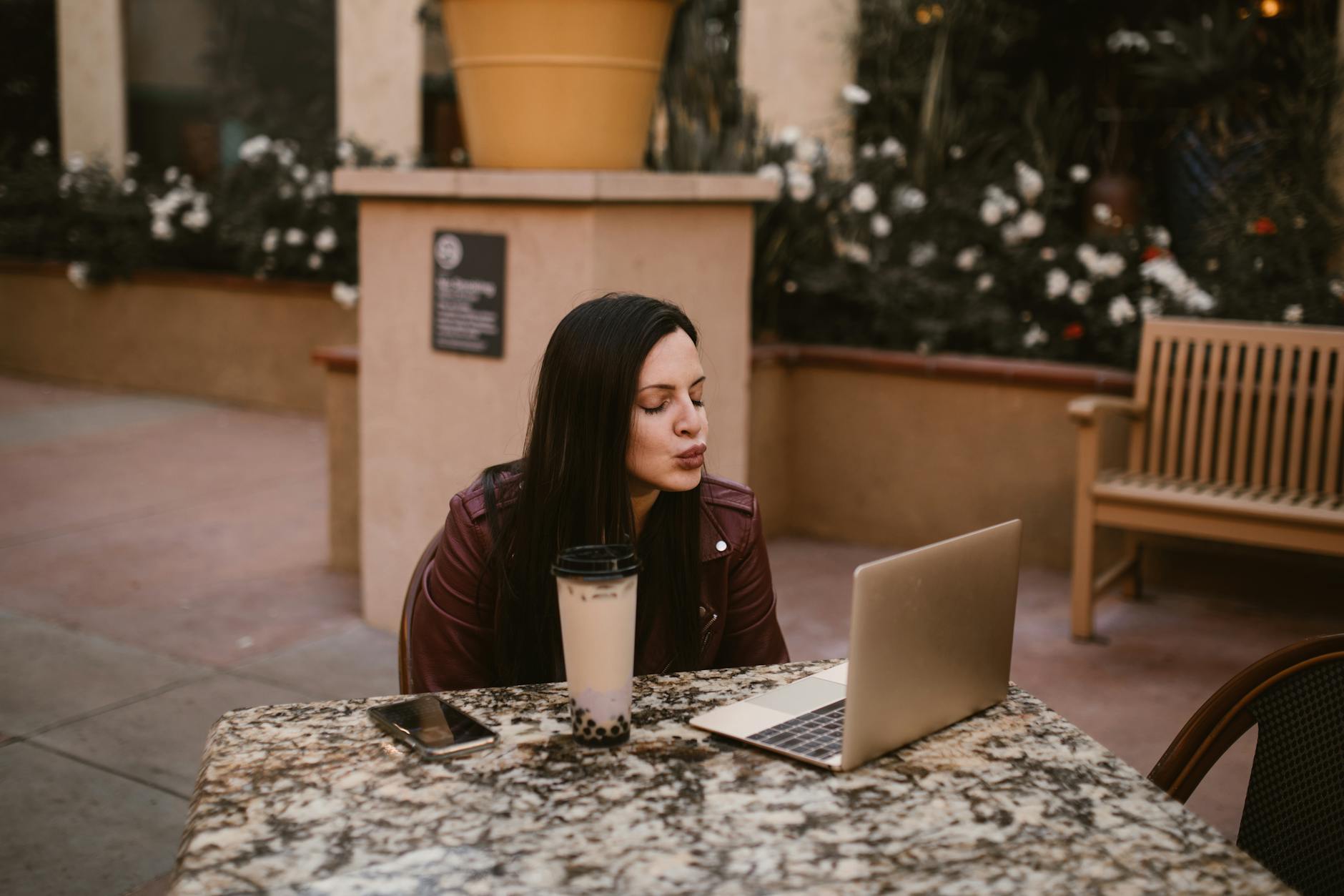 Young woman blowing a kiss during a virtual chat at an outdoor cafe table with laptop and bubble tea. - couple communication goals