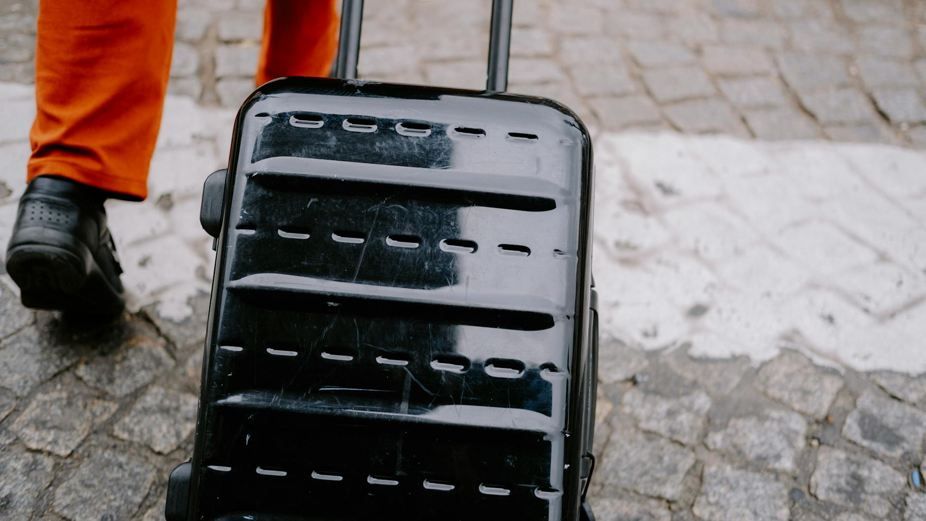 Close-up of a black suitcase being pulled on a cobblestone street by a person in orange pants. - declutter emotional baggage