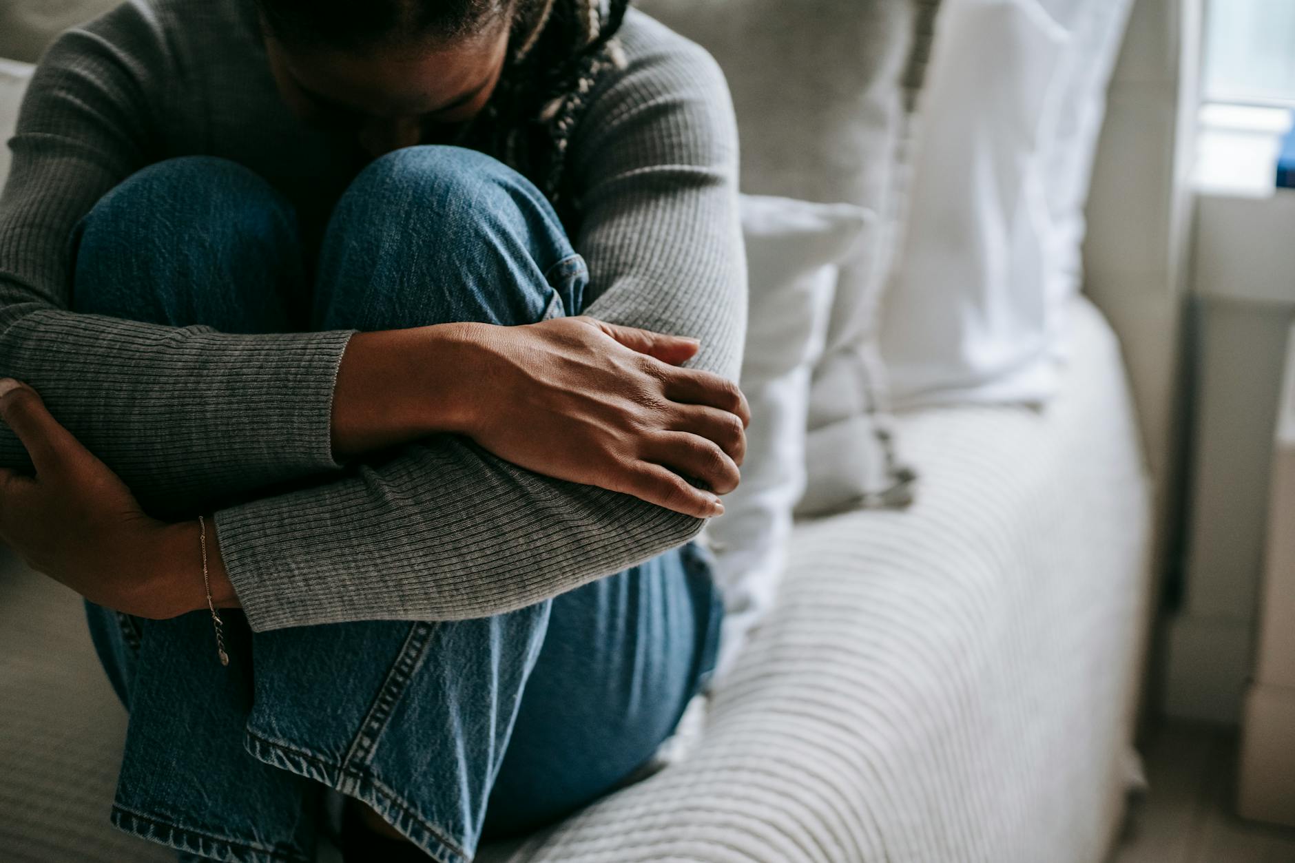 A woman sits on a bed, hugging her knees in a contemplative pose, indoors. - declutter negative thoughts