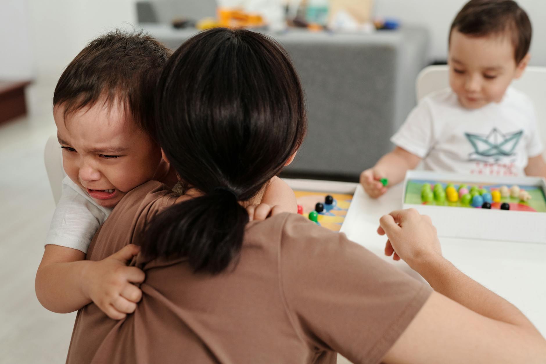 A mother comforts her crying child while another child plays nearby in a cozy home setting. - disorganized attachment
