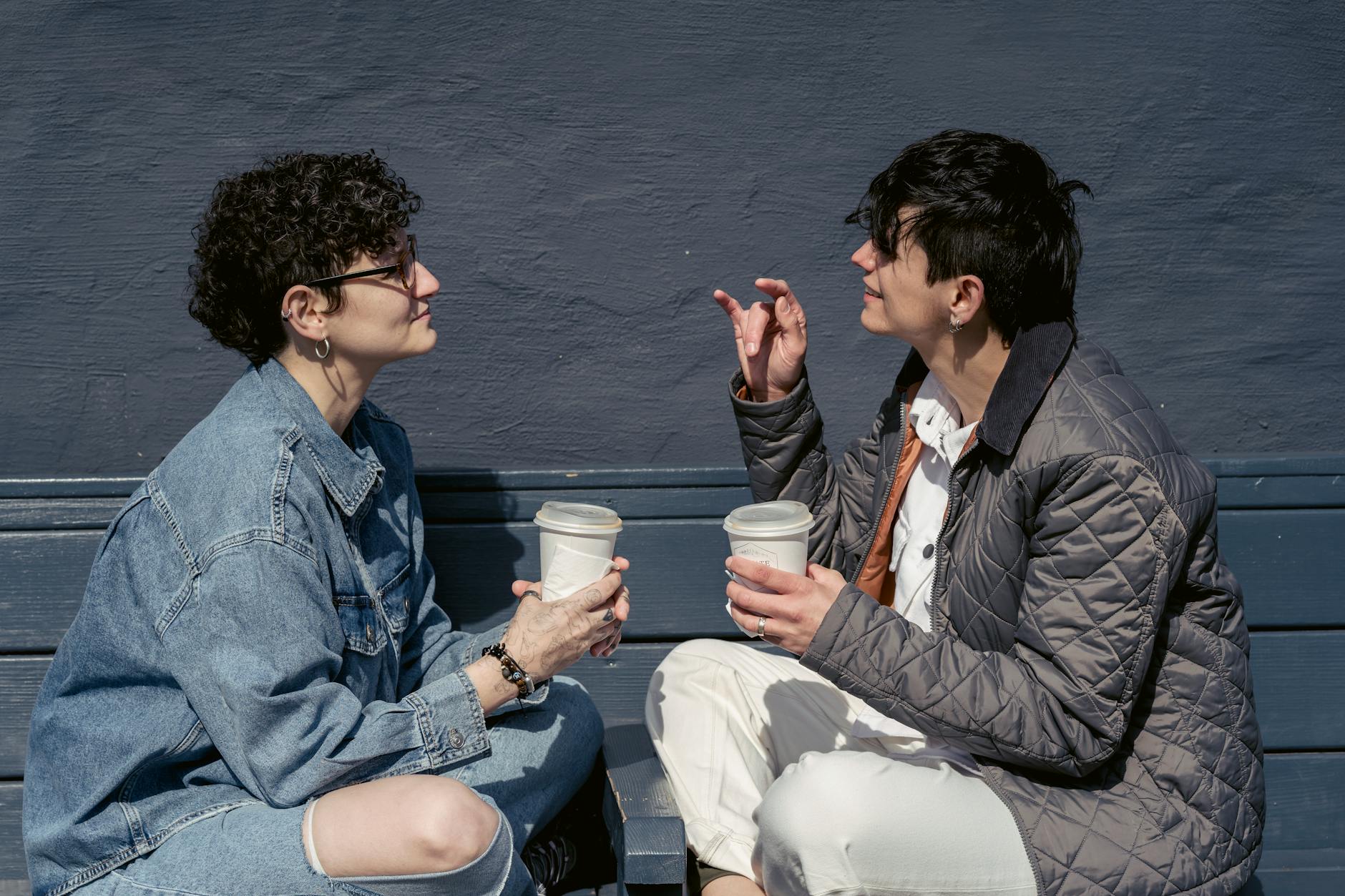 Side view of young stylish ladies with short dark hairs in casual outfits sitting on wooden bench with crossed legs and chatting while drinking takeaway coffee during weekend on street - healthy relationship communication