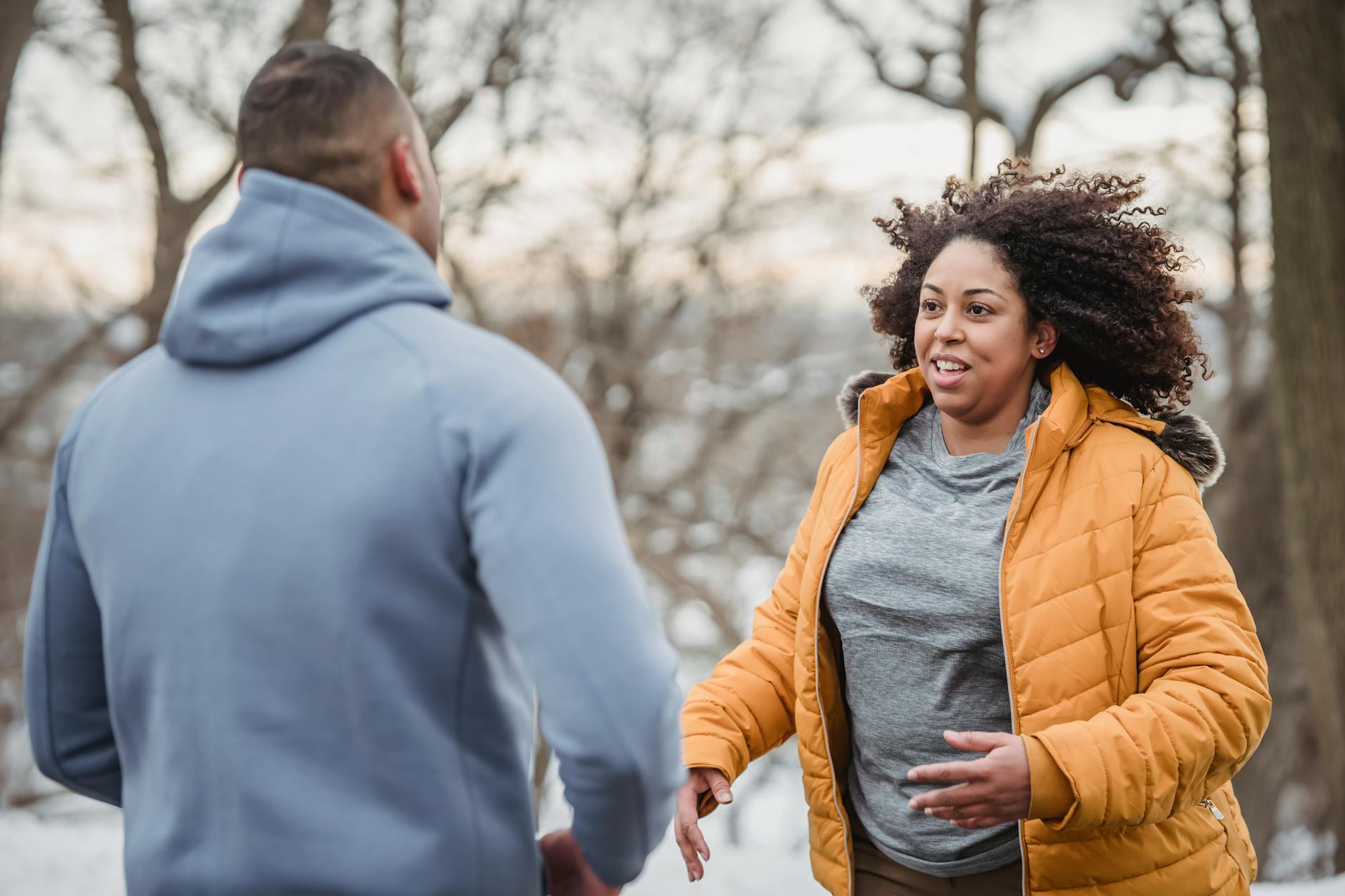 Positive plump African American in warm jacket looking at anonymous personal coach while standing in snowy nature with trees during training - healthy relationship communication