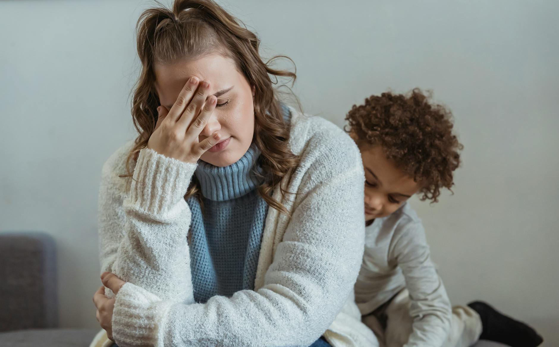 Frustrated mother with hand on forehead and closed eyes sitting near African American son near wall in room at home - parental emotional support