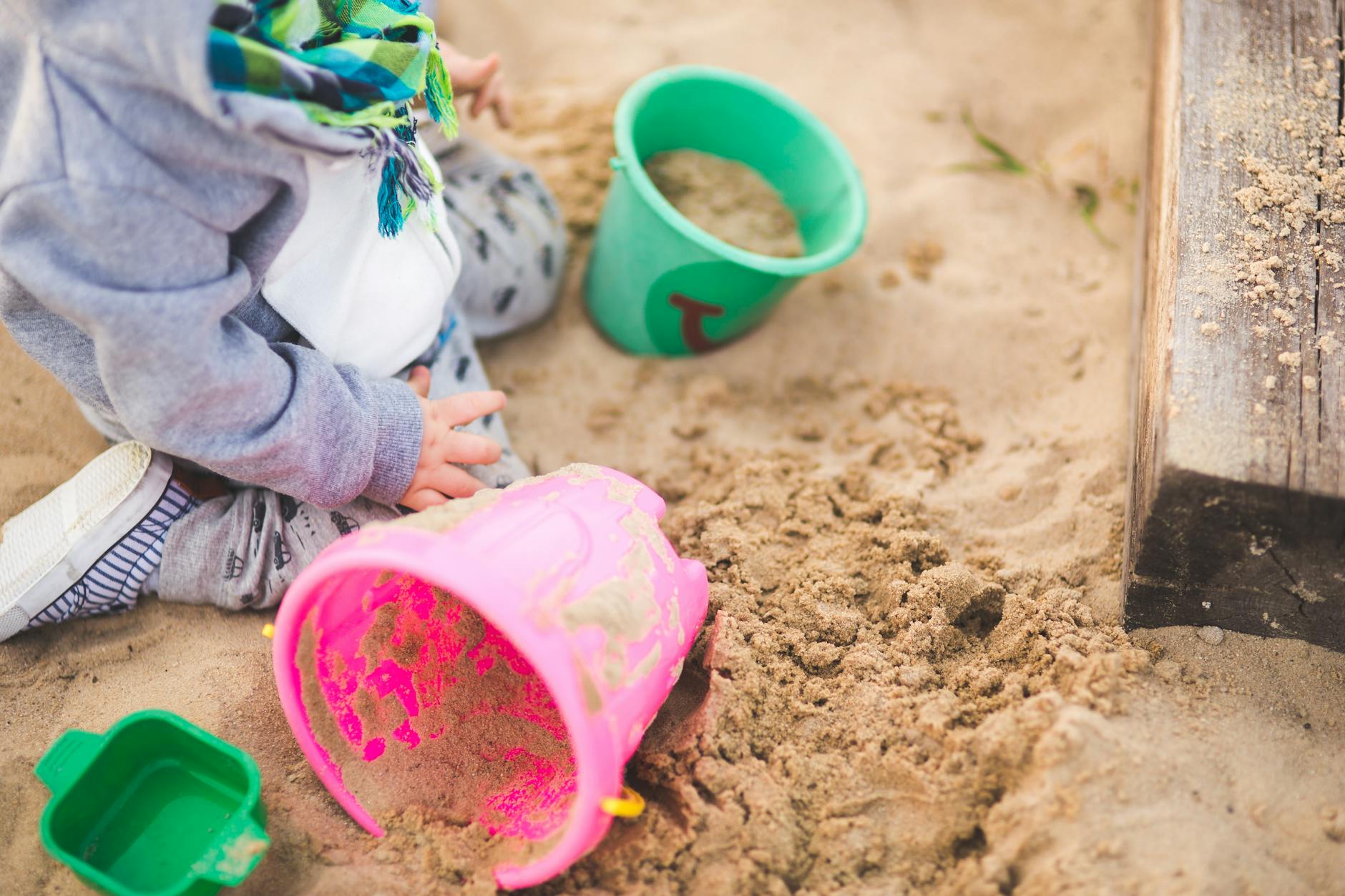 A child enjoys playing in a sandbox using vibrant colored buckets during a sunny day. - preschool social skills