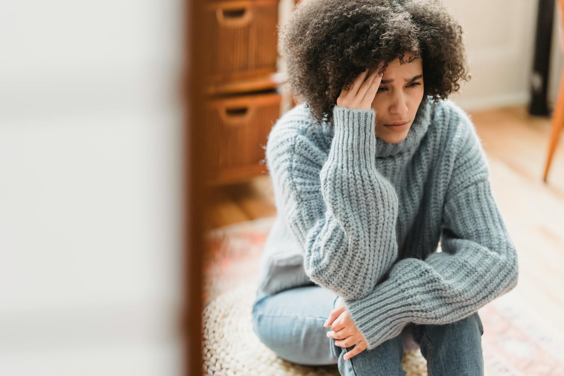 Young ethnic female in casual clothes sitting on pouf while crying with hand on head in light flat - relationship anxiety ocd