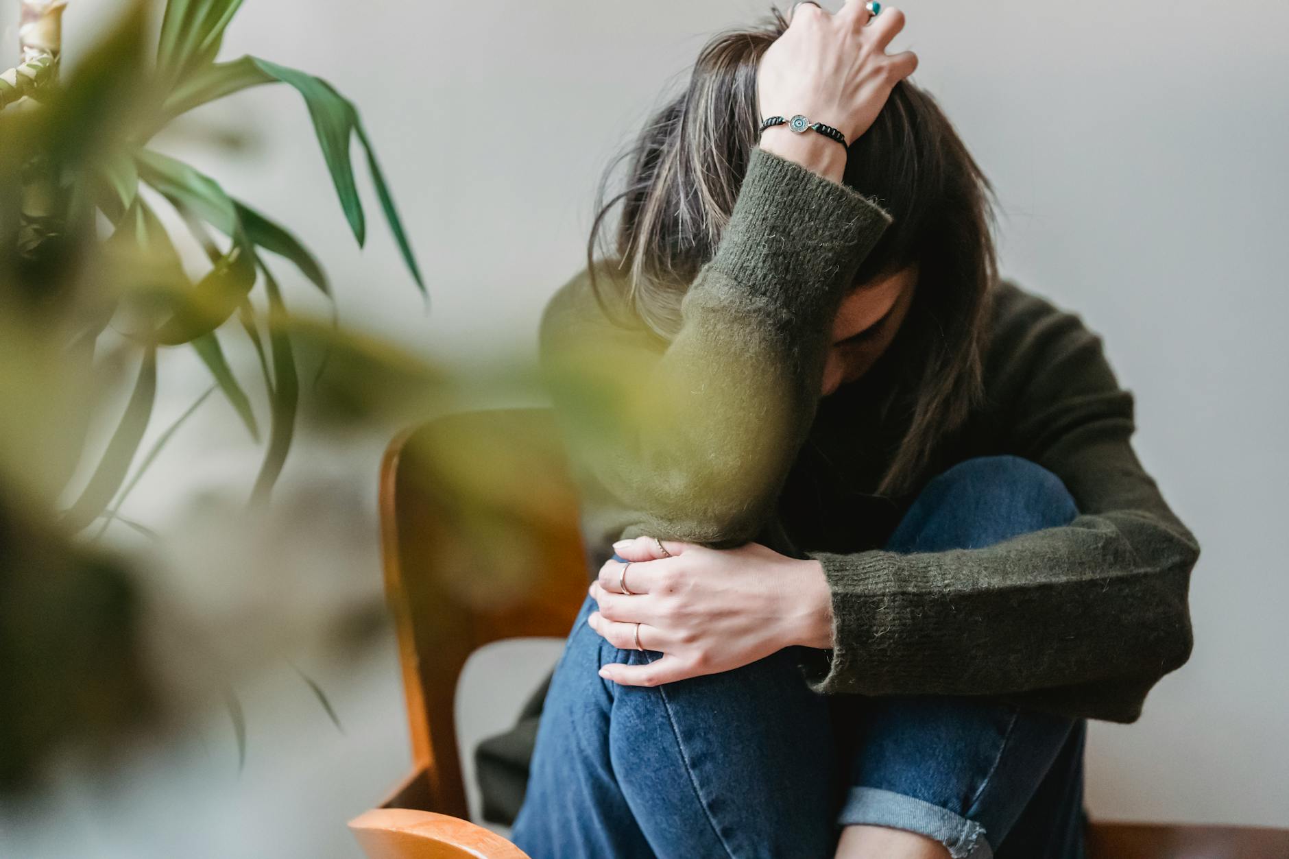 Anonymous young sorrowed female in casual outfit touching dark hair and embracing knees while sitting on chair at home - relationship anxiety ocd