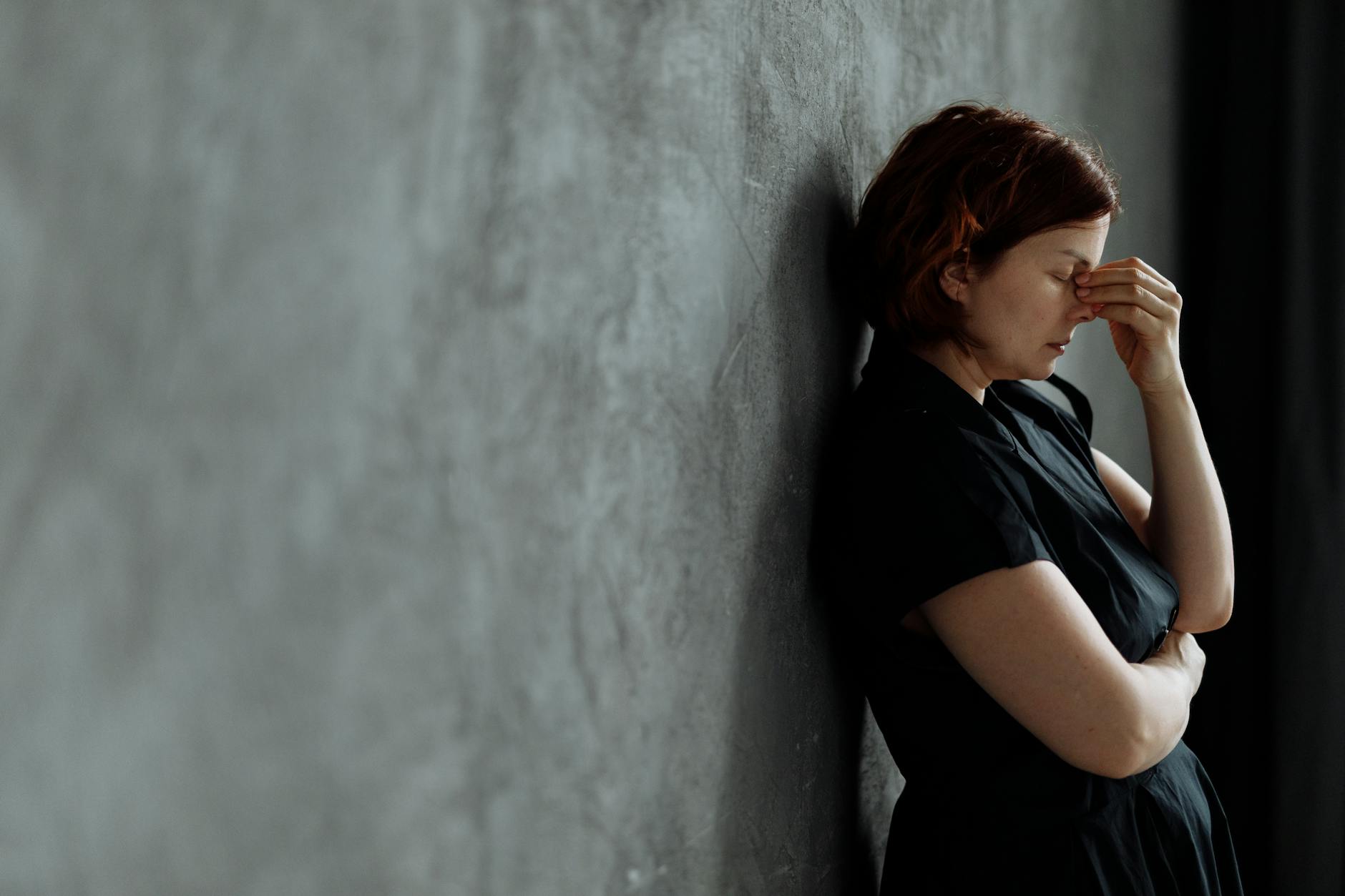 A woman in stress with eyes closed, leaning against a concrete wall indoors. - relationship anxiety reddit