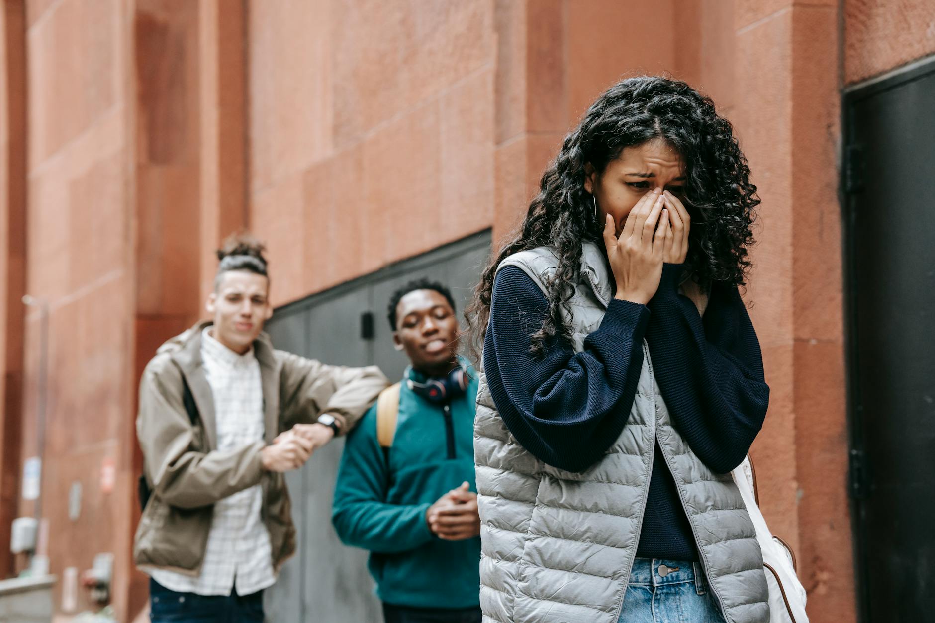 From below of despaired young ethnic female student covering mouth with hands while crying on street after being bullied by multiracial classmates - relationship anxiety spring