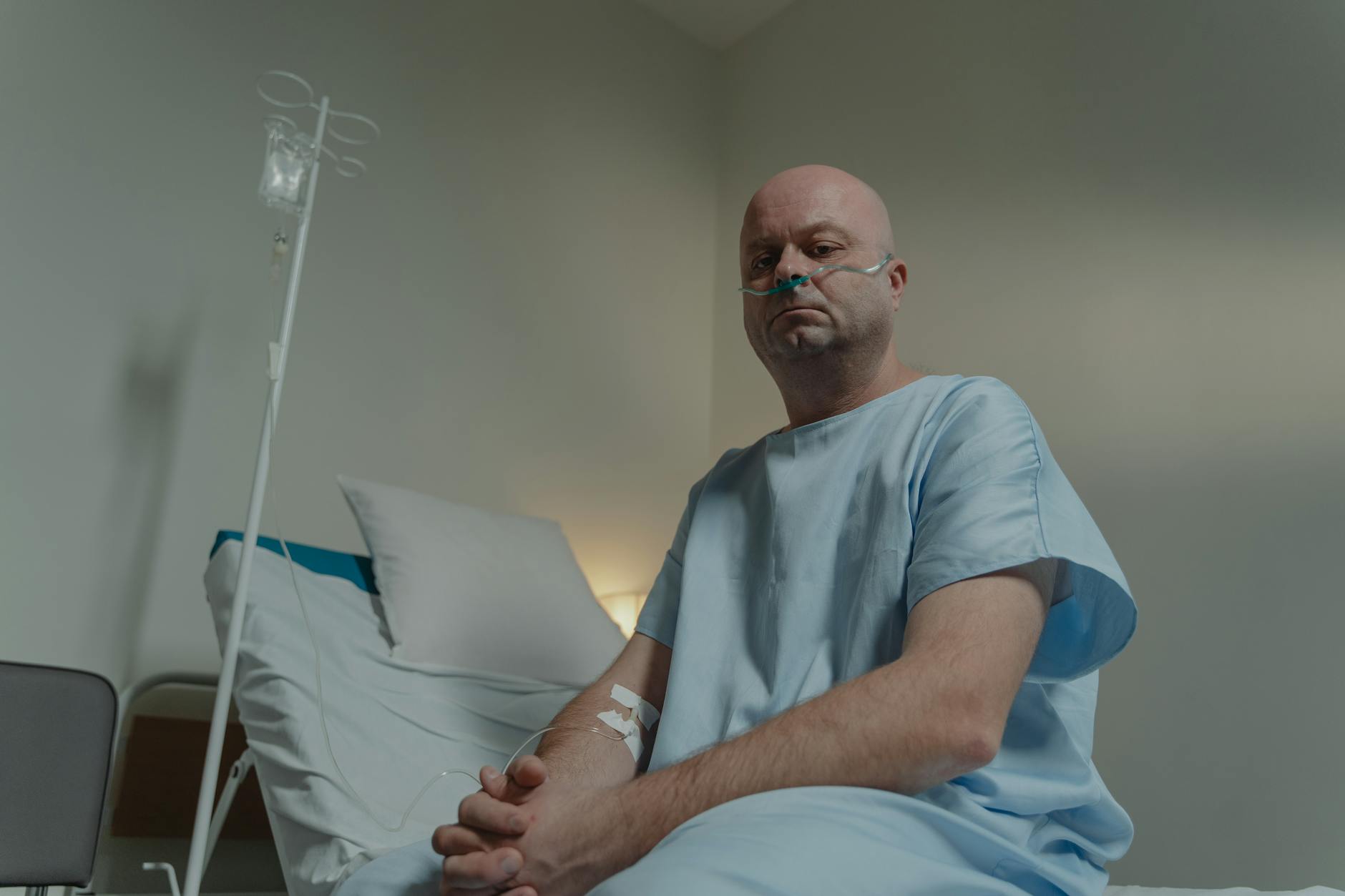 Bald man in hospital gown sitting on bed with nasal cannula and IV in a clinical setting. - sad spring treatment